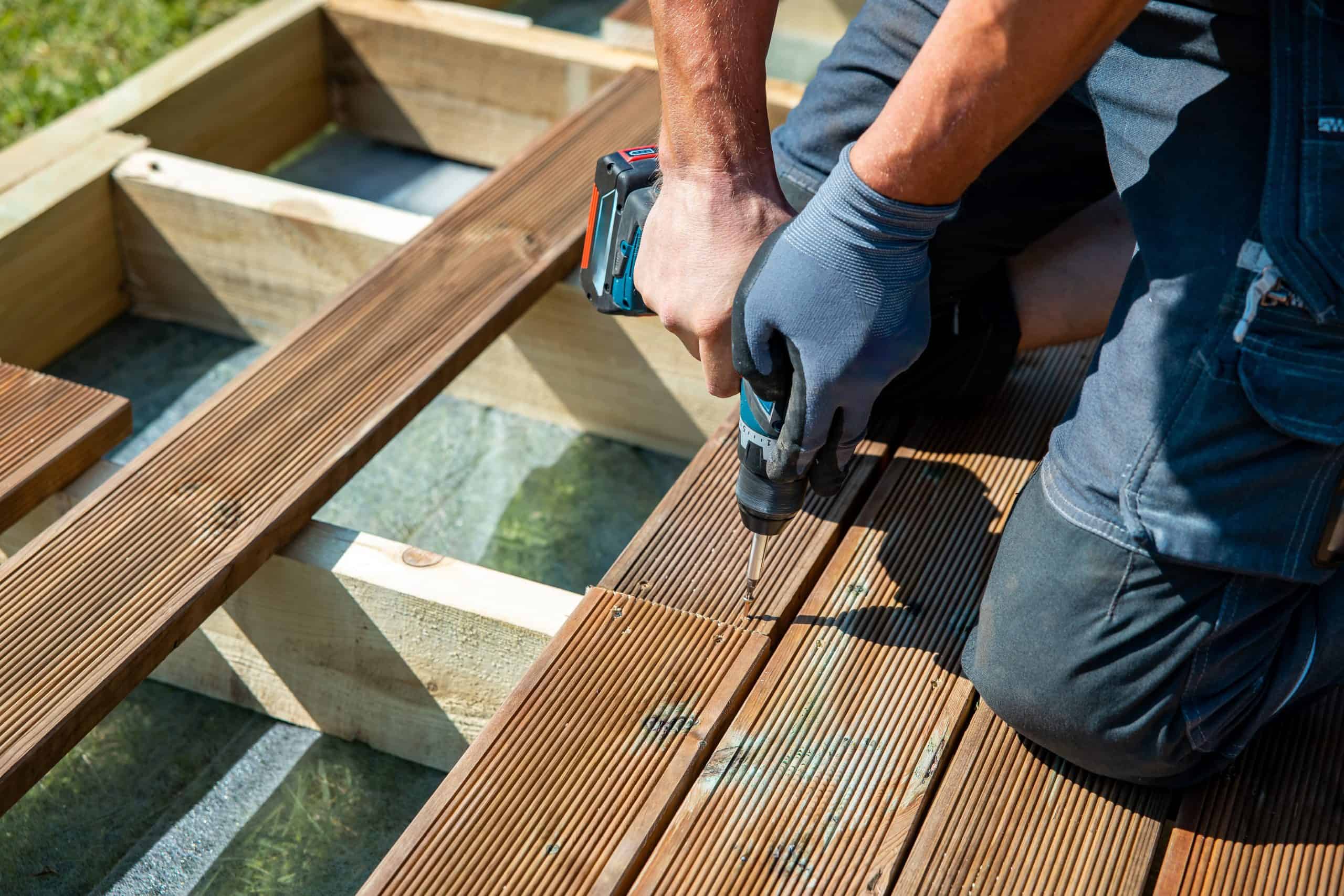 A person wearing gloves is using a cordless drill to install wooden decking under the warm sunlight, casting shadows on the grooved, brown boards.