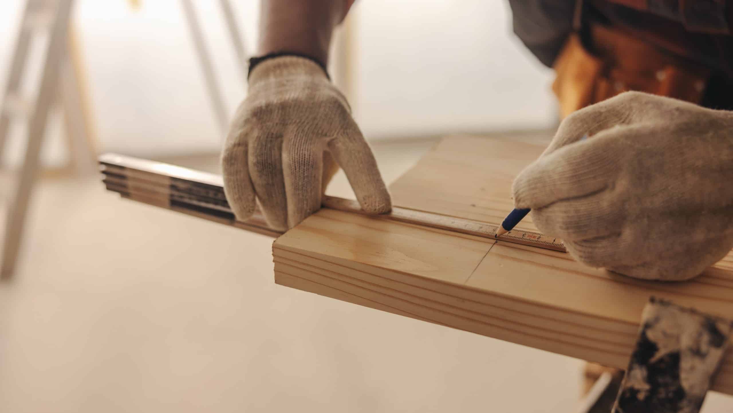 Close-up of a person wearing gloves using a pencil to mark a measurement on a wooden board with the aid of a ruler.