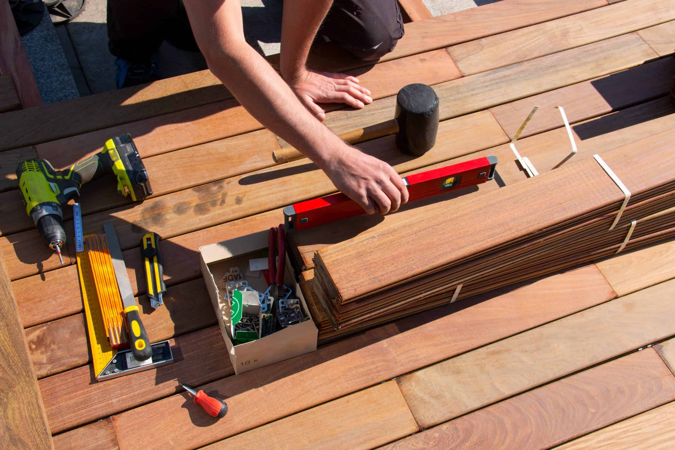 A person meticulously arranges wooden planks on a deck, surrounded by tools like a drill, mallet, and screws.