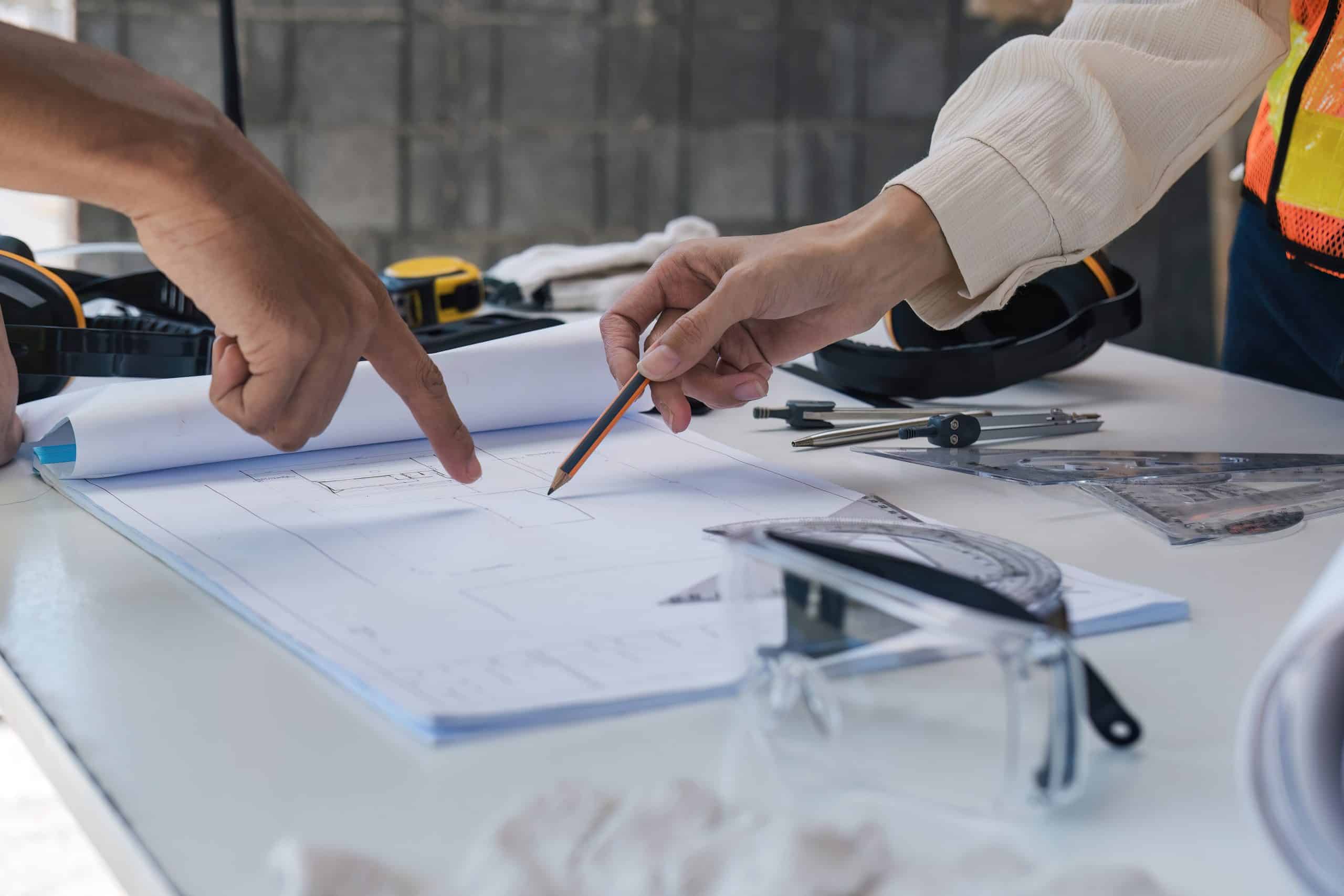 Two people are pointing at a set of blueprints on a table laden with drafting tools like compasses and rulers.
