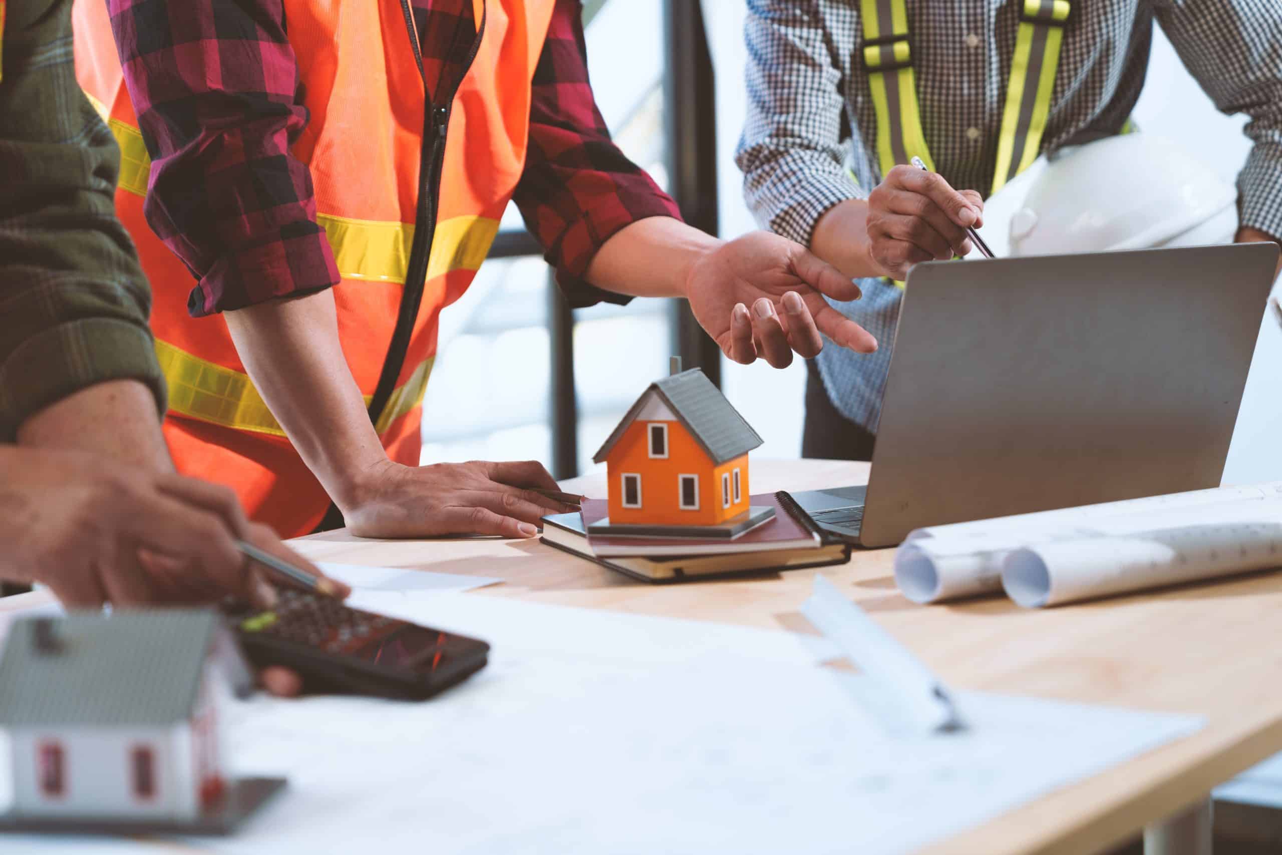 Three construction workers discuss plans at a table with blueprints, a laptop, and a small house model.