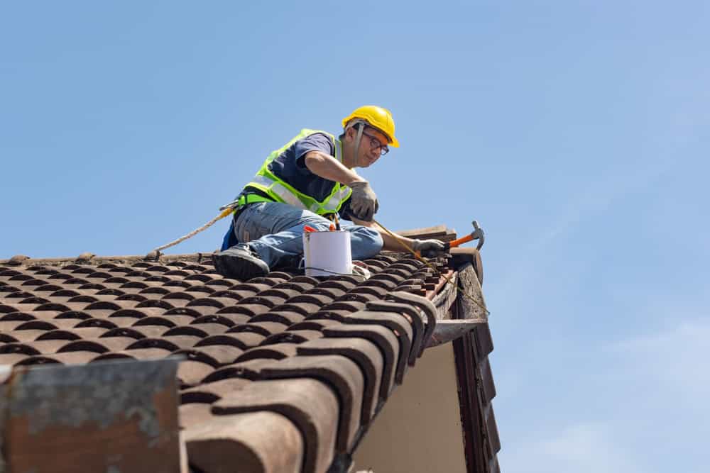 A construction worker in a yellow hard hat and green safety vest repairs a rooftop with brown tiles under a clear blue sky.