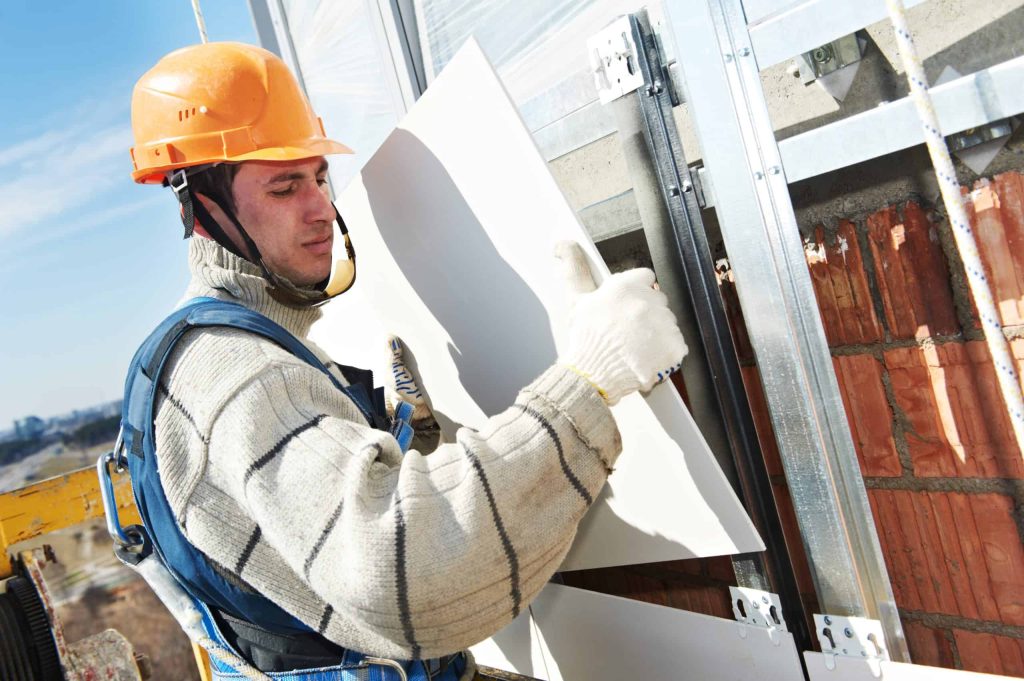 A construction worker in protective gear, including a hard hat and gloves, installs a panel on a building exterior.