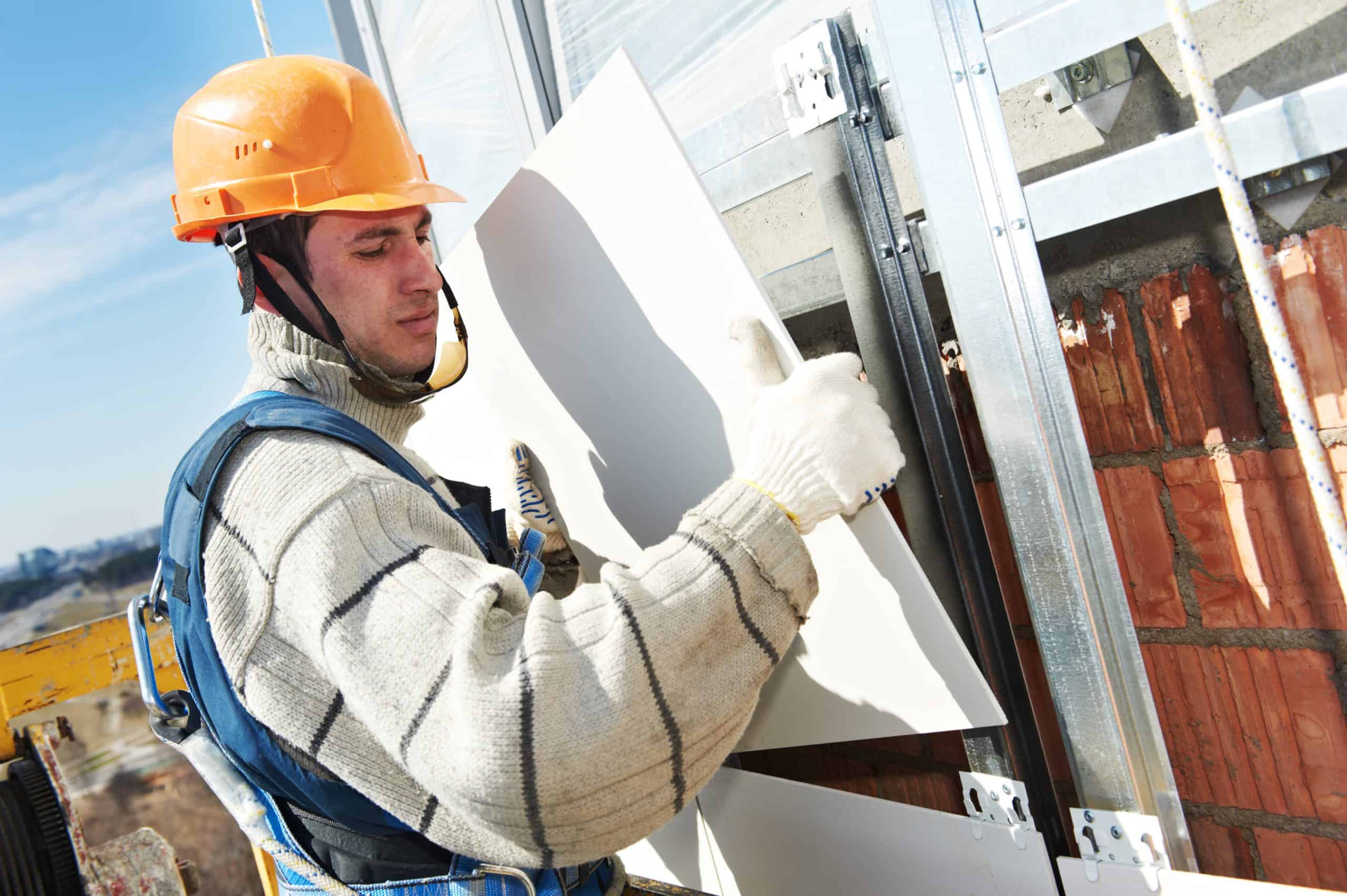 A construction worker in a striped sweater and overalls installs a panel on a brick wall, skillfully working on the building's exterior.