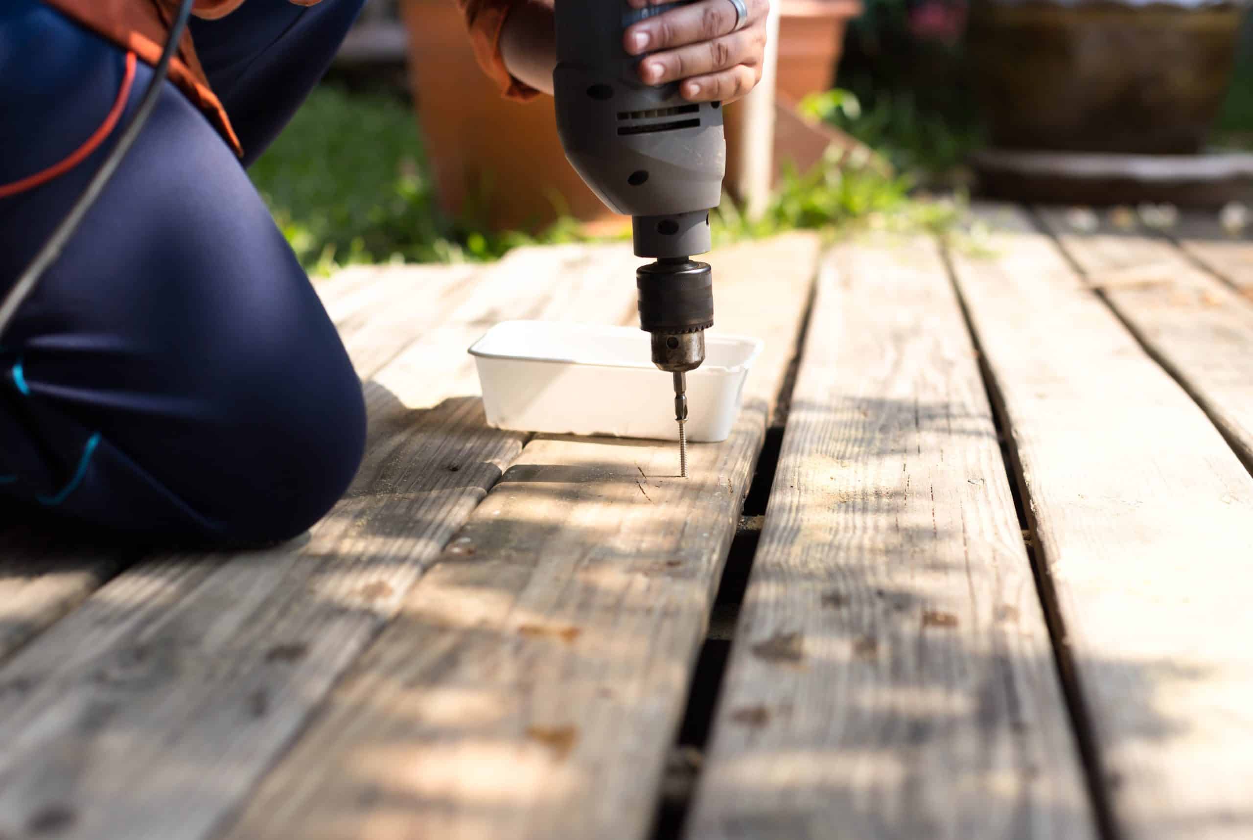 A person kneels on a wooden deck, using a power drill to secure a fastener into place.