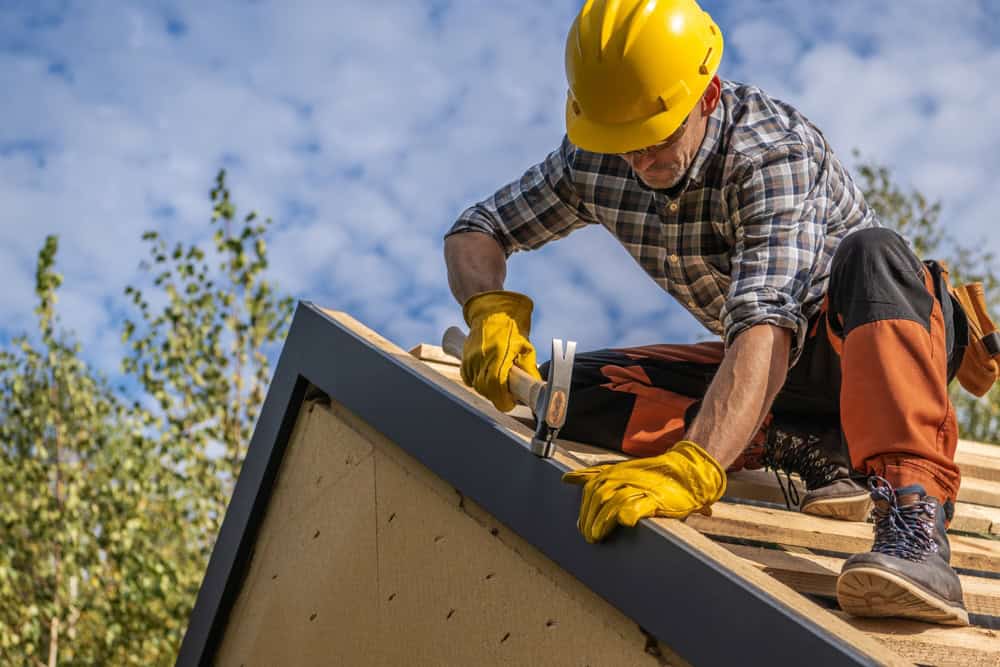 A construction worker in a yellow hard hat, gloves, and safety gear kneels on a roof’s edge, hammering diligently.