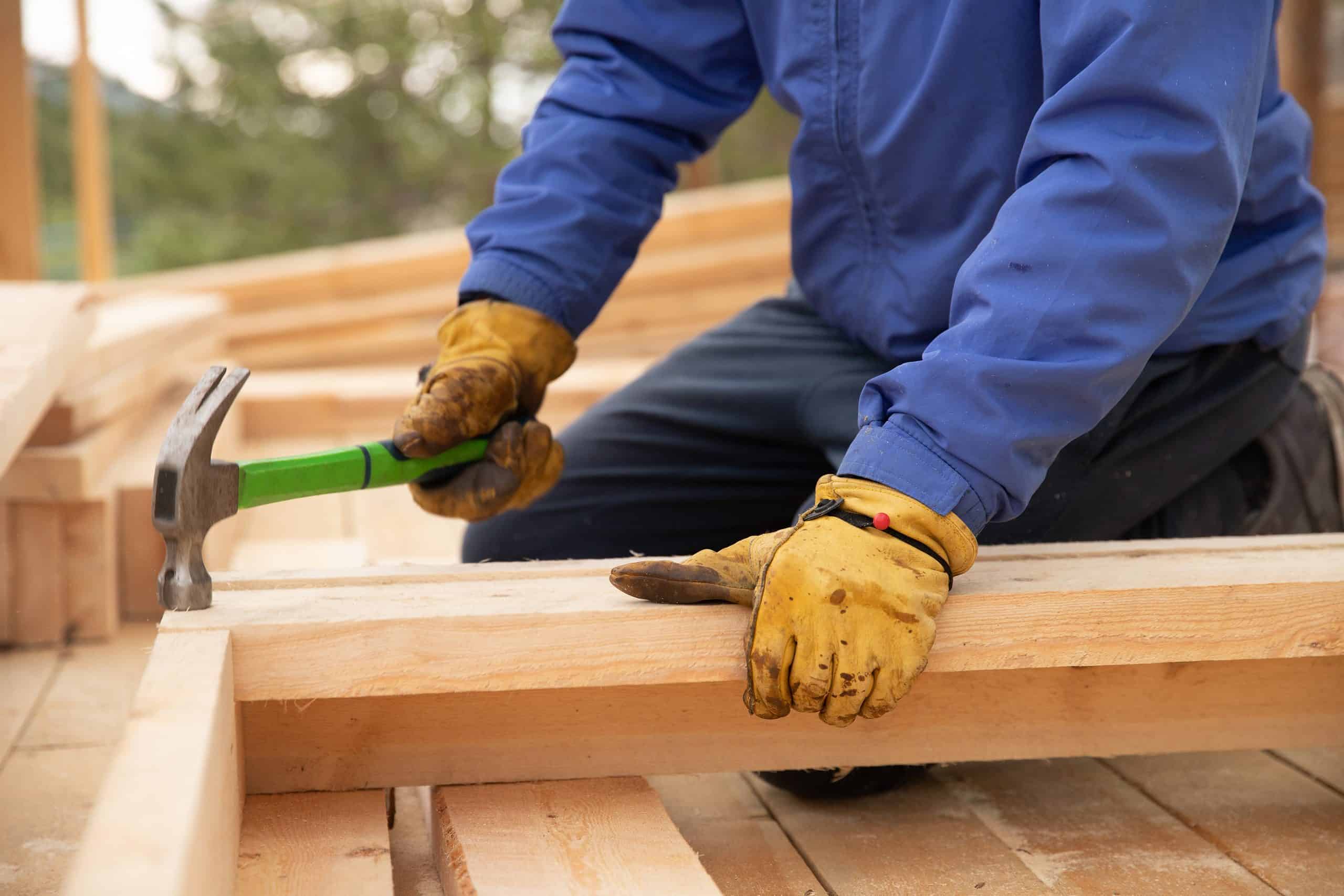 A person wearing a blue jacket and yellow gloves hammers two wooden beams together at a construction site.