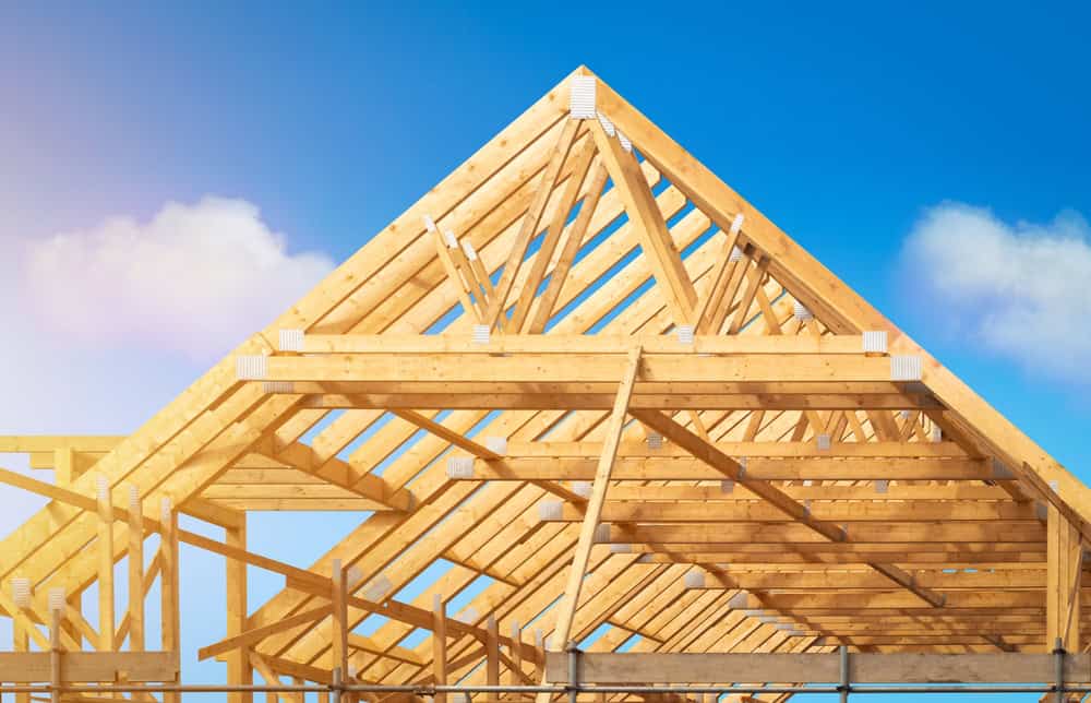 A wooden house frame under construction gleams under the bright blue sky with a few clouds, as sunlight bathes the beams in a warm, golden hue.