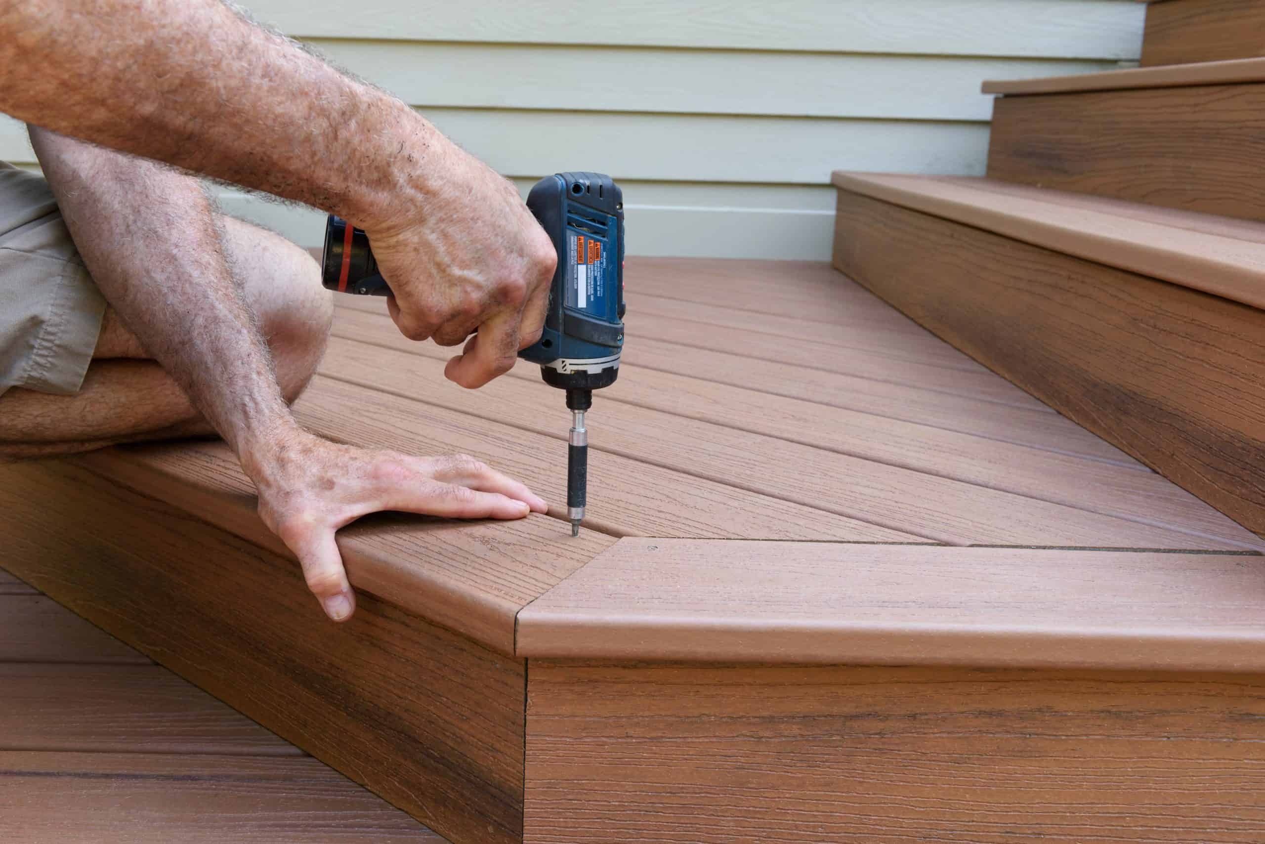 A person using a power drill to secure a brown composite deck board on an outdoor staircase.