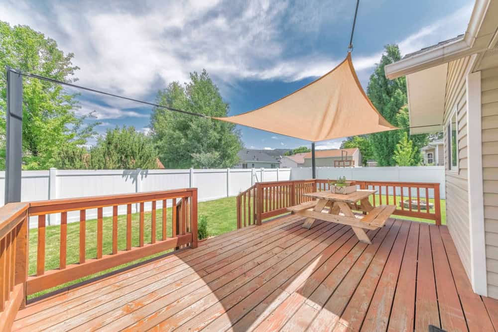 Sunny backyard with a wooden deck and picnic table beneath a triangular shade sail.