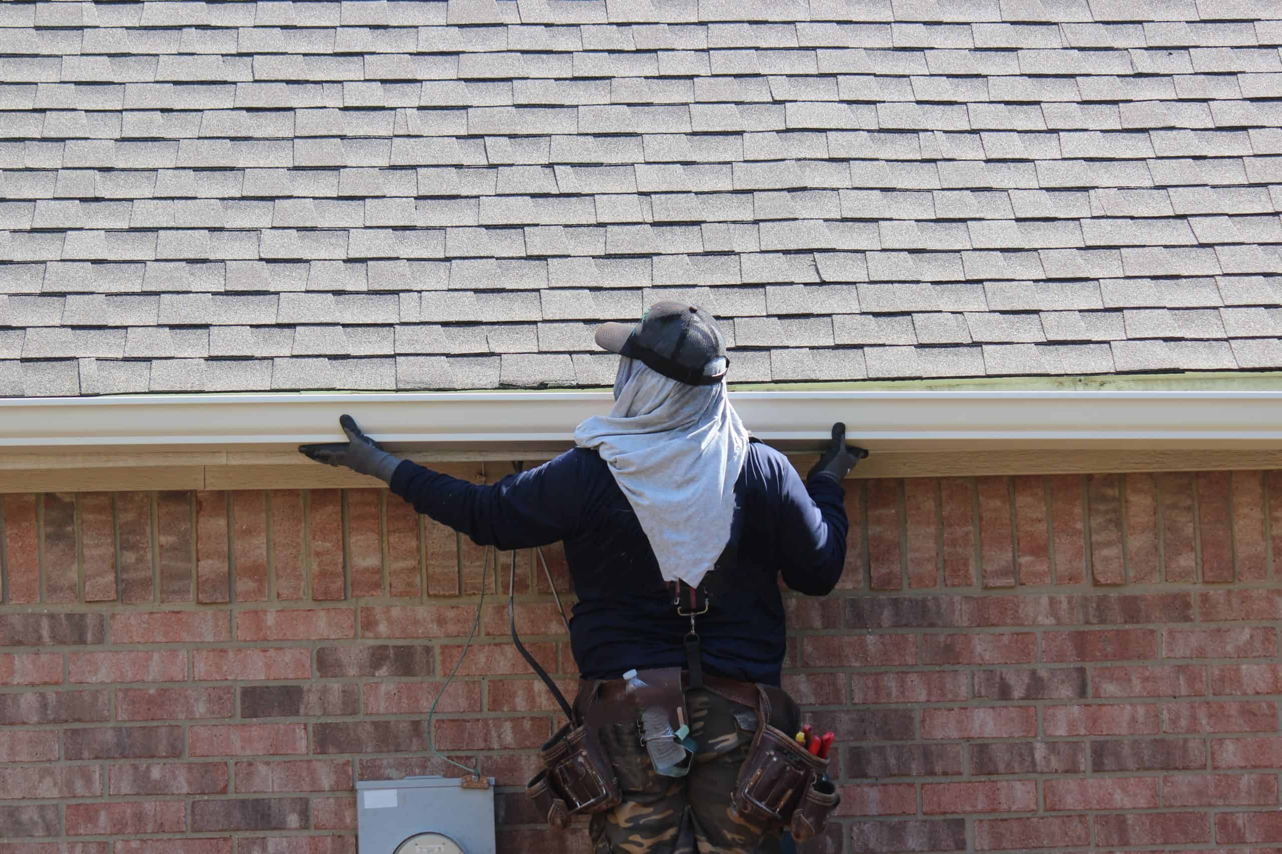 A person wearing a cap, long-sleeve shirt, and work gloves is adjusting a gutter on a brick house.