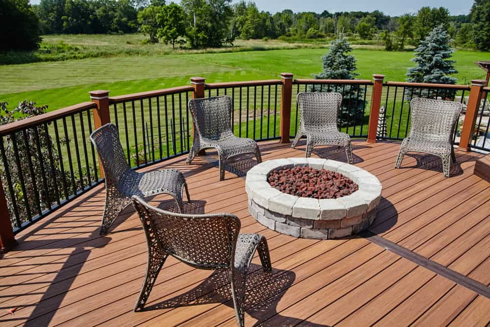 Five wicker chairs are arranged in a semi-circle around a stone fire pit on a wooden deck.
