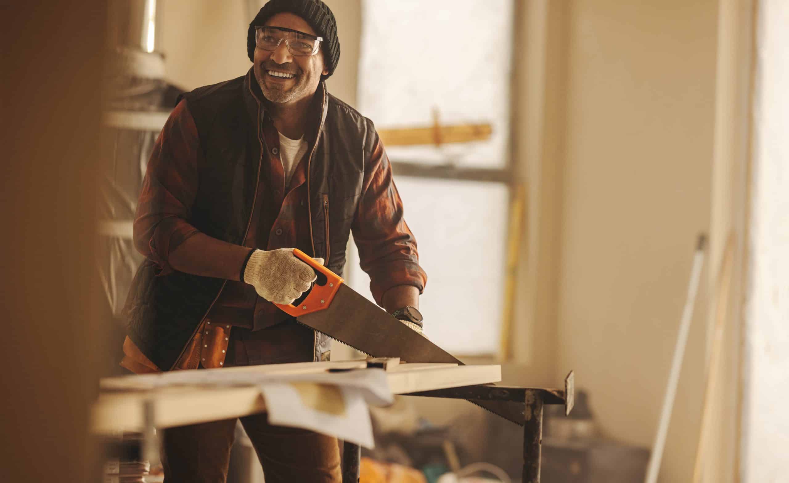 A man wearing a beanie, safety glasses, and gloves is smiling while using a handsaw to cut a piece of wood in his workshop.