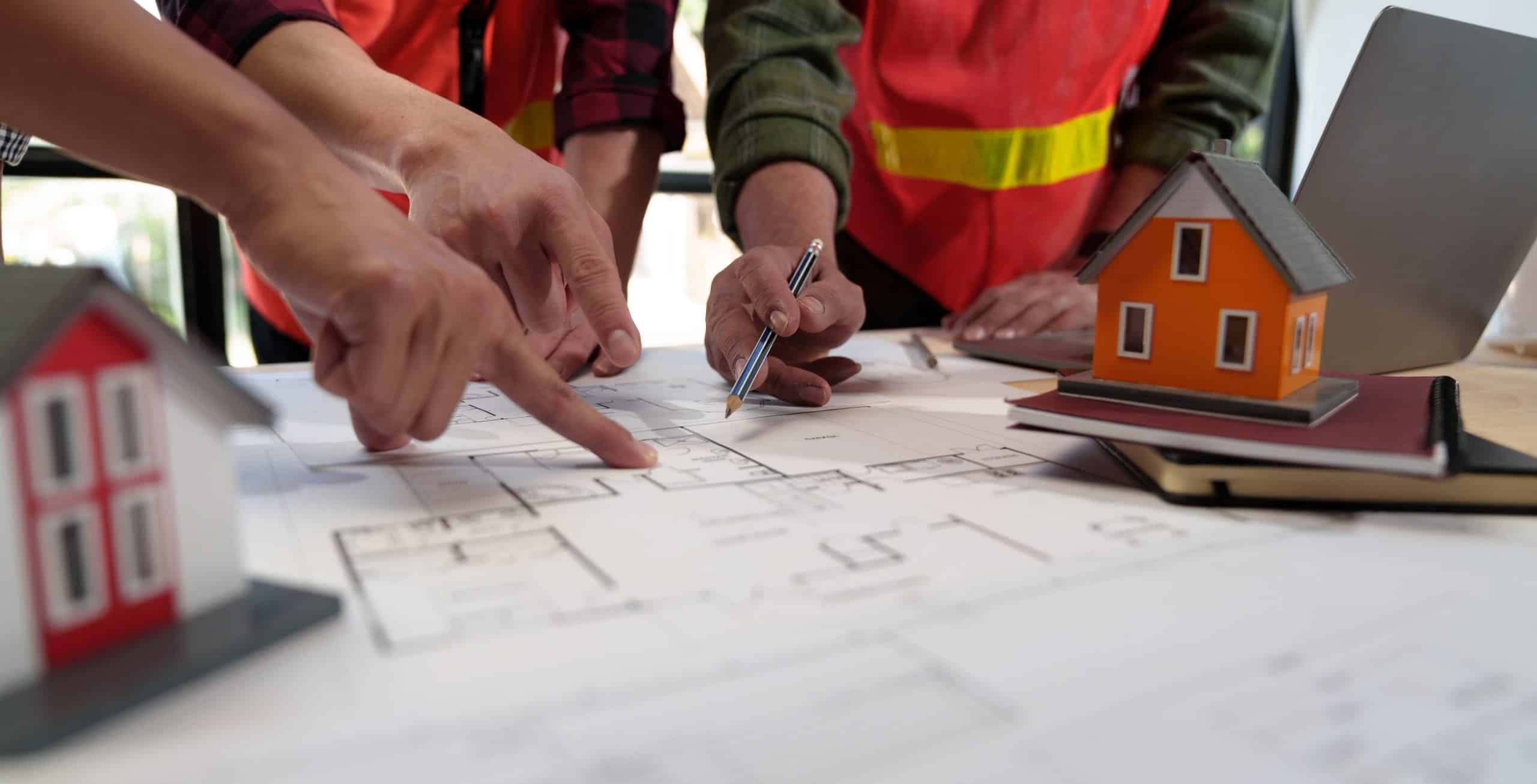People in reflective vests discuss architectural blueprints on a table, pointing at the plans with a pen.