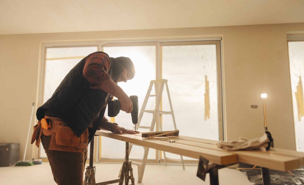 A person in a beanie hat uses a power drill on wooden planks placed on a sawhorse in a sunlit room, showcasing their home remodeling skills.