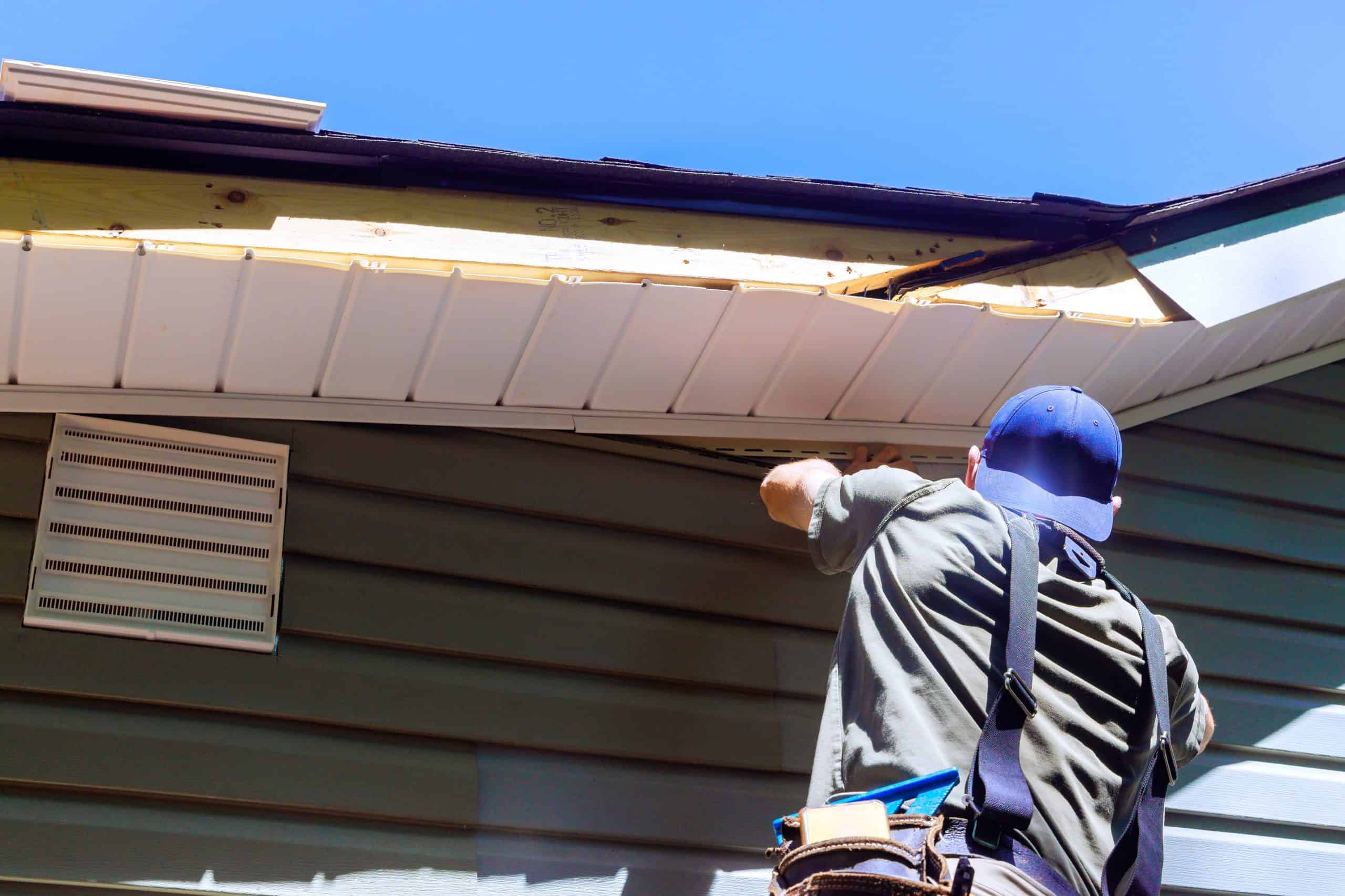 A person in a blue cap and gray shirt is diligently working on the siding installation of a house, adjusting a piece near the unfinished roof edge that exposes the wood.