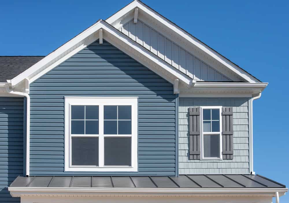Close-up of a beautifully remodeled house exterior against a clear blue sky, featuring a large white-framed window and a smaller side window with grey shutters.