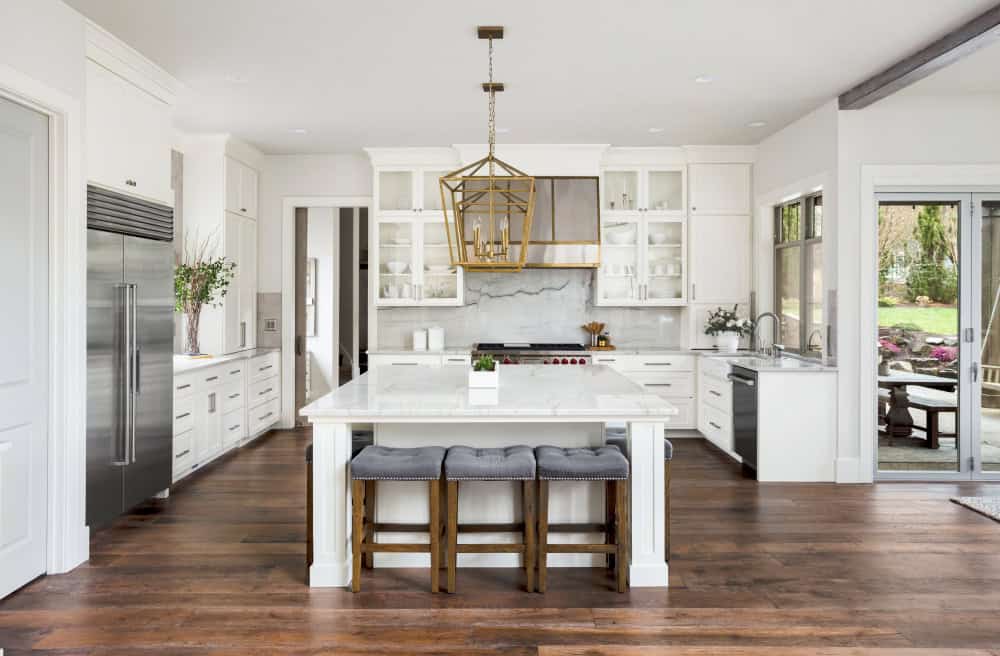 A modern kitchen with white cabinets, a marble island, and gray cushioned stools. A geometric pendant light hangs above the island.