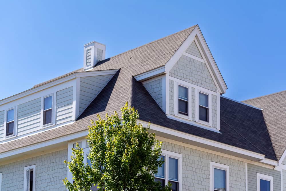 A light grey house, recently remodeled by a general contractor in Orange County, NY, features multiple windows and a shingled roof set against a clear blue sky. A leafy green tree partially obscures the modern, angular architecture.