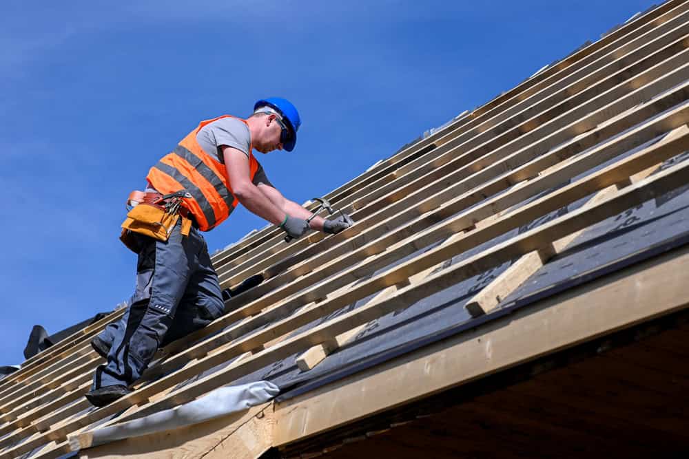 A construction worker in a blue helmet and orange safety vest is busy on a sloped roof under a clear blue sky, expertly using a hammer to secure wooden beams. As tools and materials surround them, they carry out precise tasks fit for home remodeling in Orange County, NY.