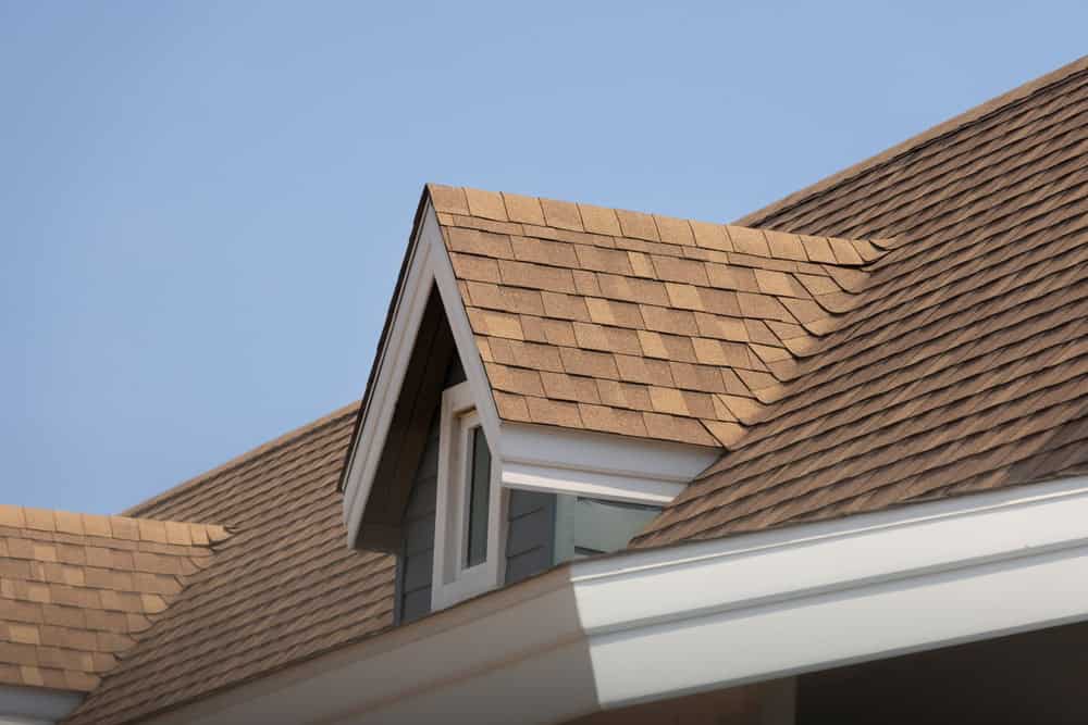 A close-up view of a house roof with brown shingles, featuring a gable dormer and a small window, ready for roof inspections in Dutchess County NY. The sky in the background is clear and blue.
