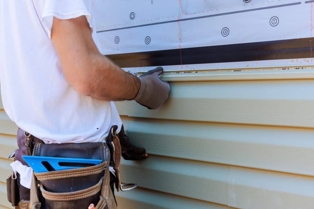 A person wearing a white t-shirt and brown tool belt is expertly installing green vinyl siding on a building in Dutchess County, NY.