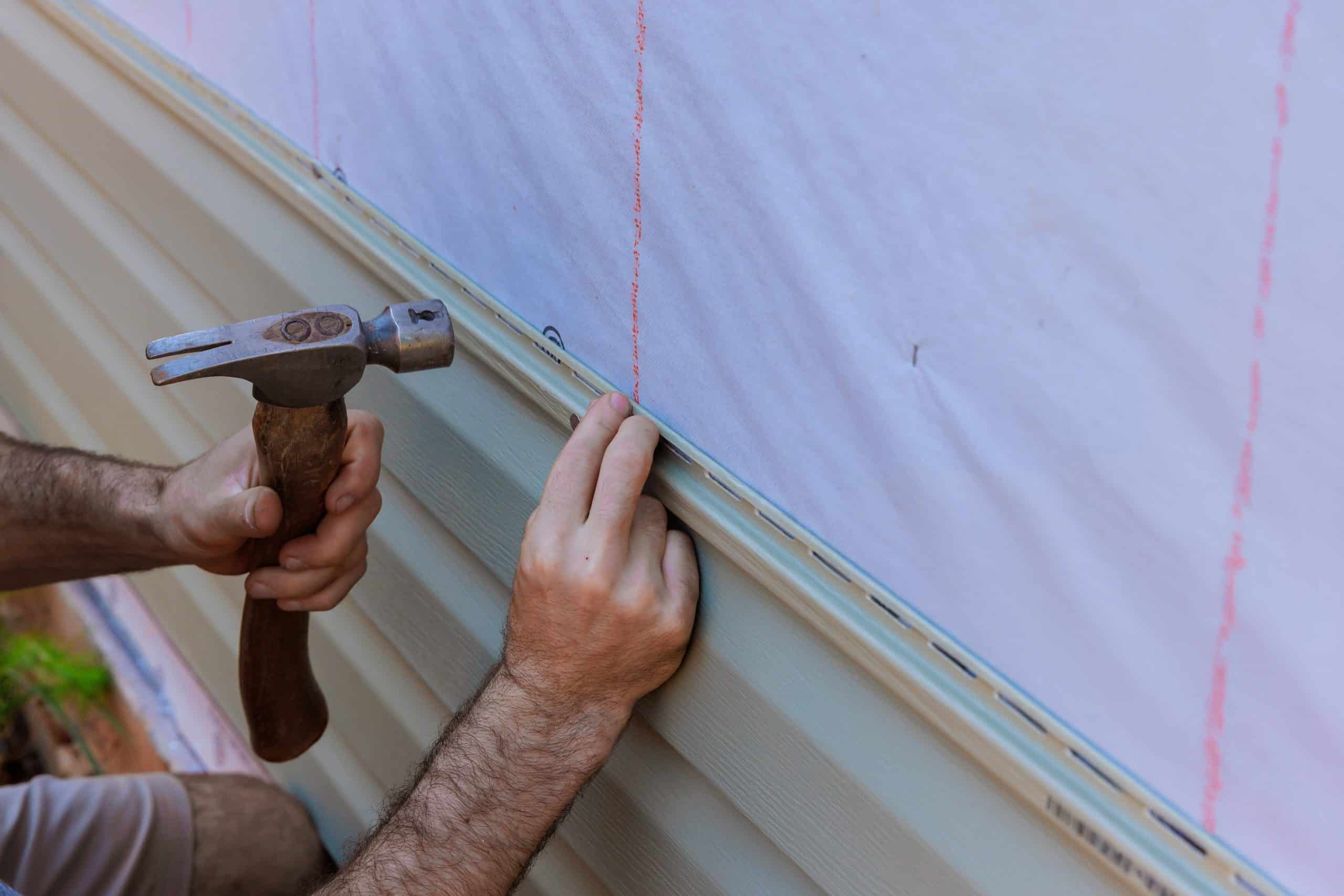A general contractor in Orange County, NY, is securing beige vinyl siding to a home's exterior wall using a hammer. The wall features a white underlayment with red guidelines. The contractor skillfully holds the material steady while completing the siding installation.