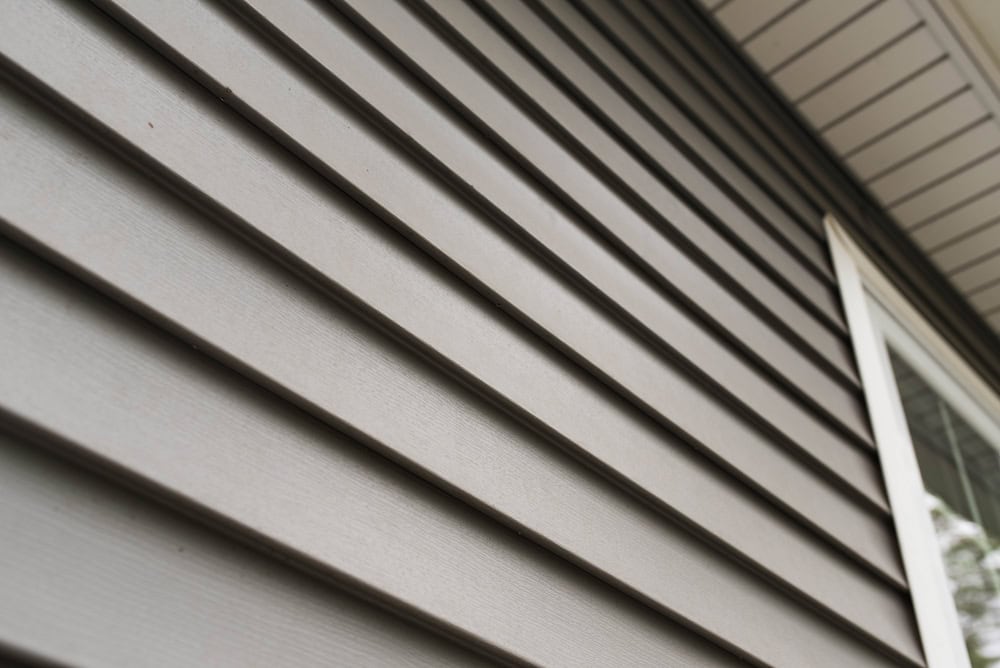 Close-up of a house exterior in Dutchess County, NY, featuring expertly installed horizontal gray siding and a partial view of a white-trimmed window on the right. The angle emphasizes the texture and lines of the siding, with a portion of the roof's soffit visible at the top.
