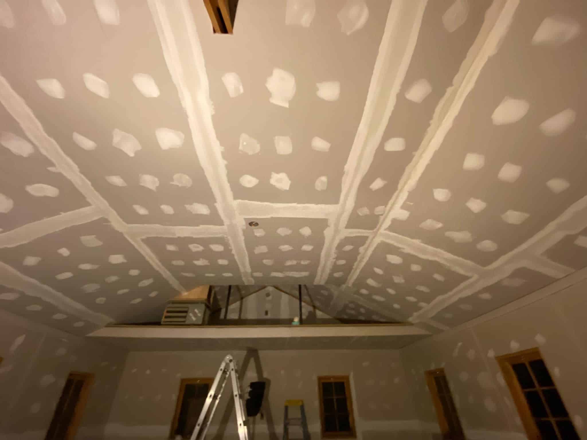 A room with unfinished drywall on the ceiling. Various tools and ladders are positioned around the room. The ceiling has visible joint compound on seams and screw spots. Natural wooden window frames are present on the walls.
