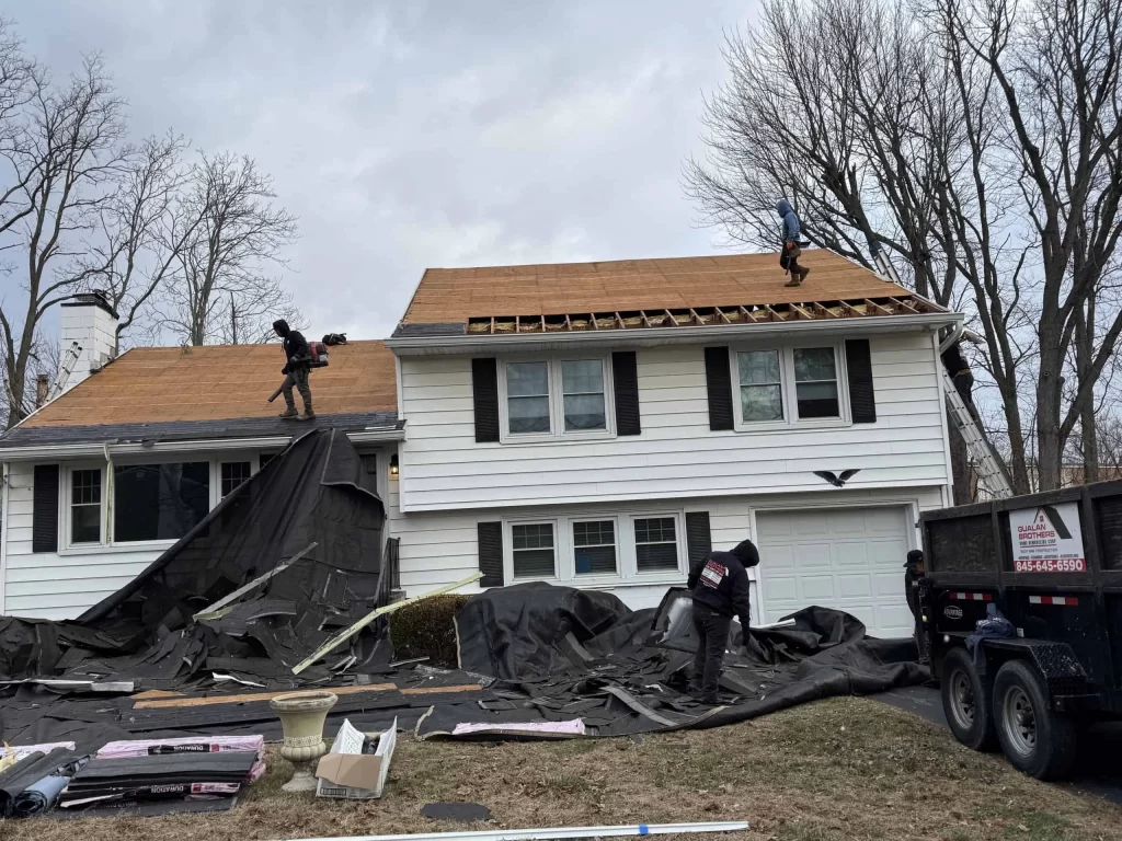 Workers are repairing the roof of a two-story white house with black shutters.
