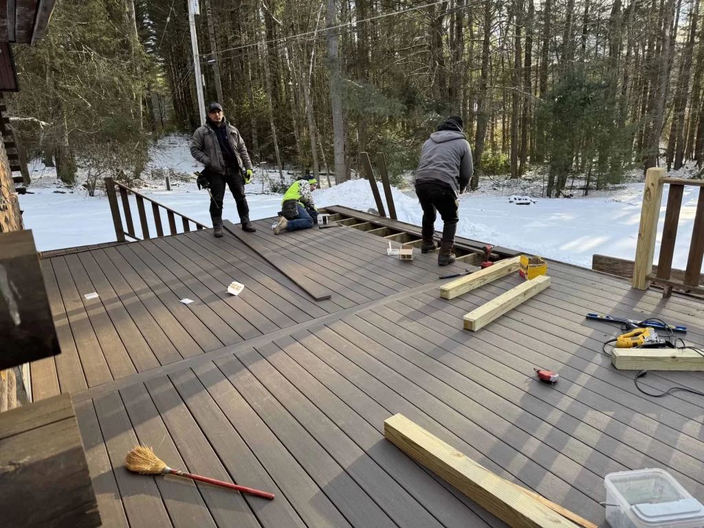 Three people work on constructing a wooden deck in a snowy forest setting.