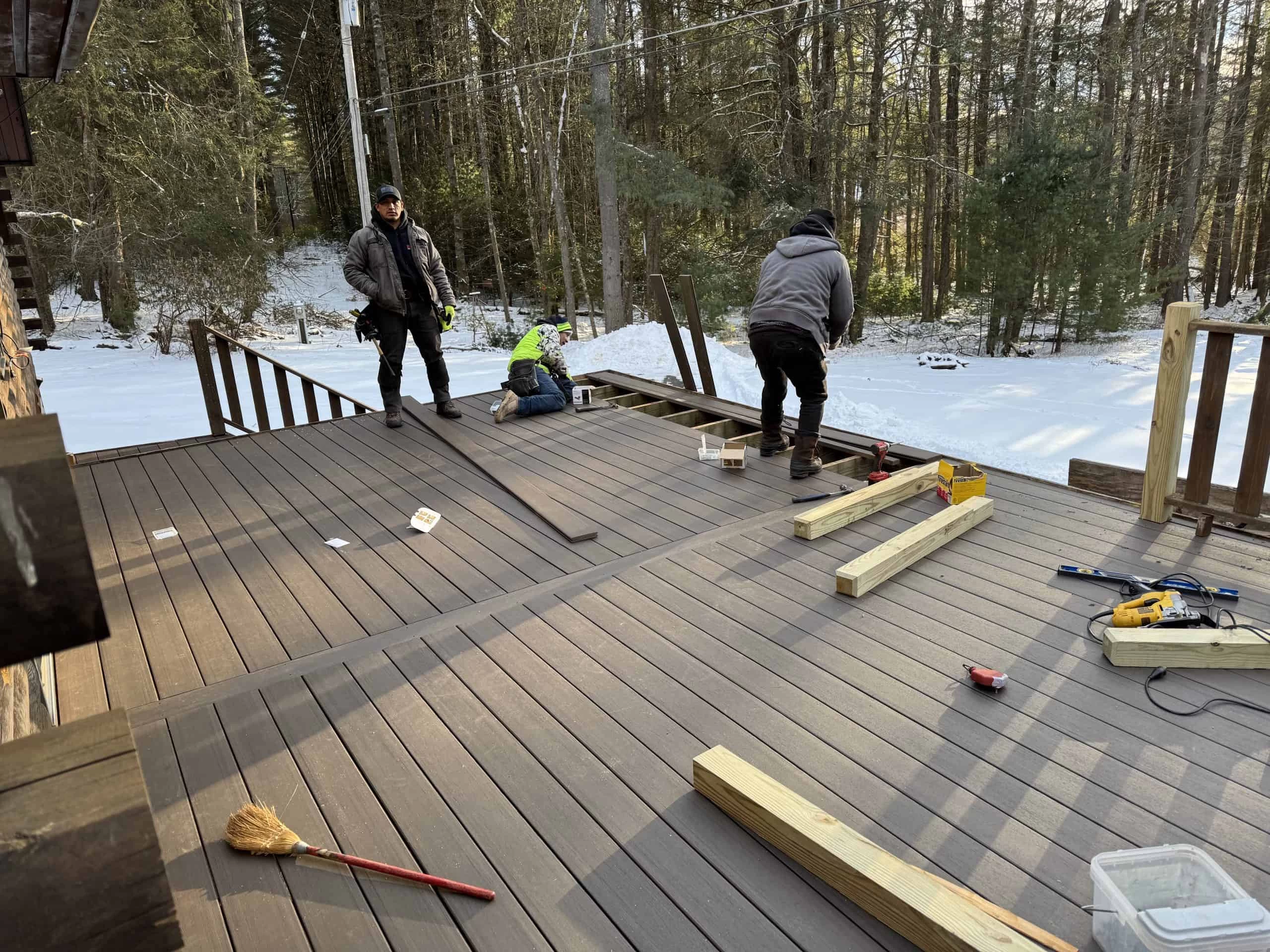 Three people work on constructing a wooden deck in a snowy forest setting.