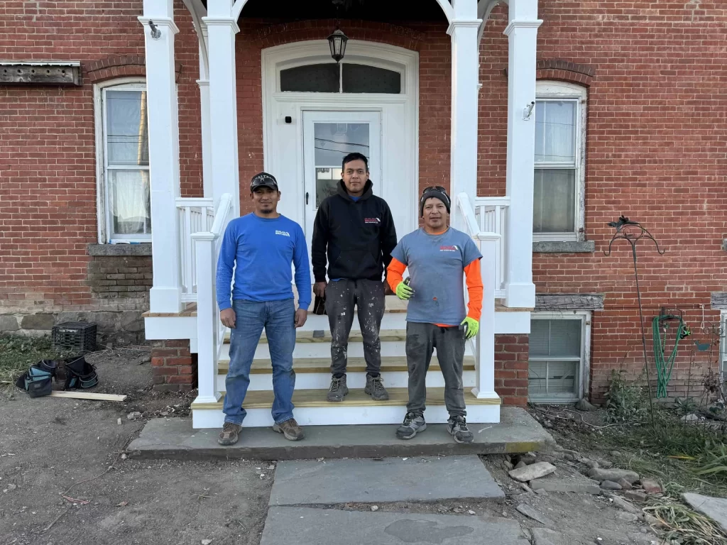 Three construction workers stand on the steps of a red brick building with a white door and porch.