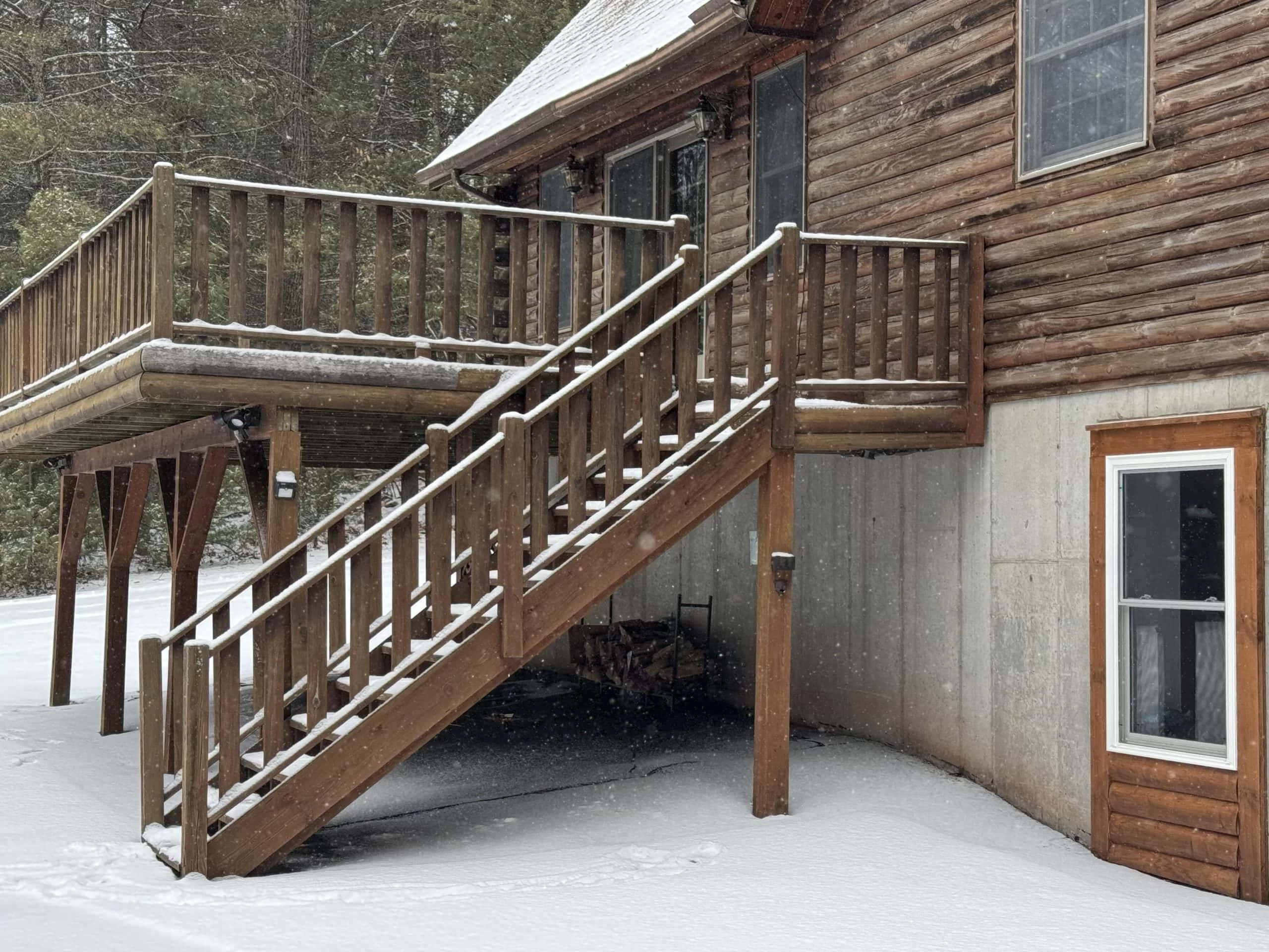 A wooden staircase leads up to a raised deck attached to a log cabin in a snowy landscape.