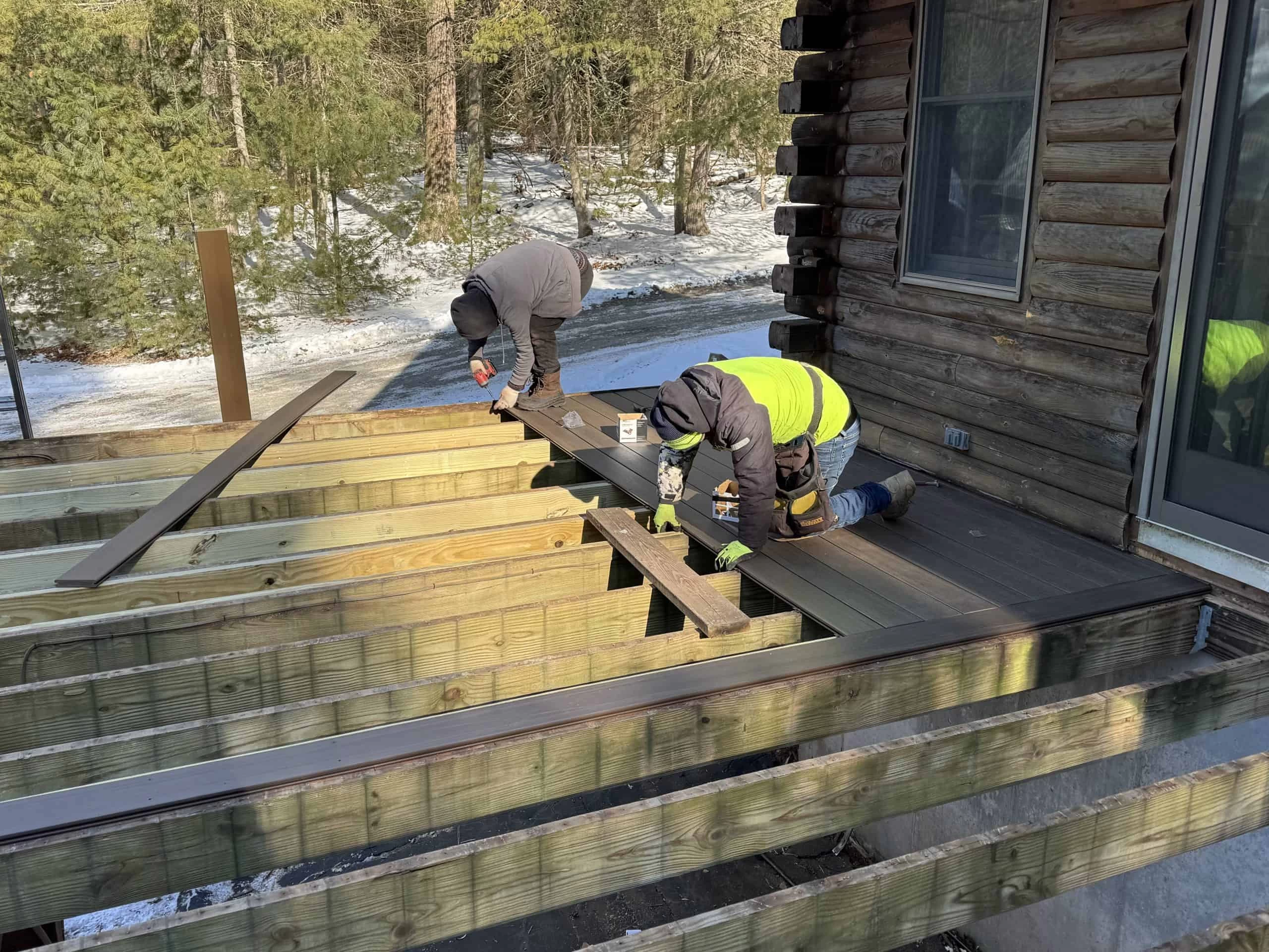 Two workers build a wooden deck attached to a log cabin in a snowy outdoor setting.