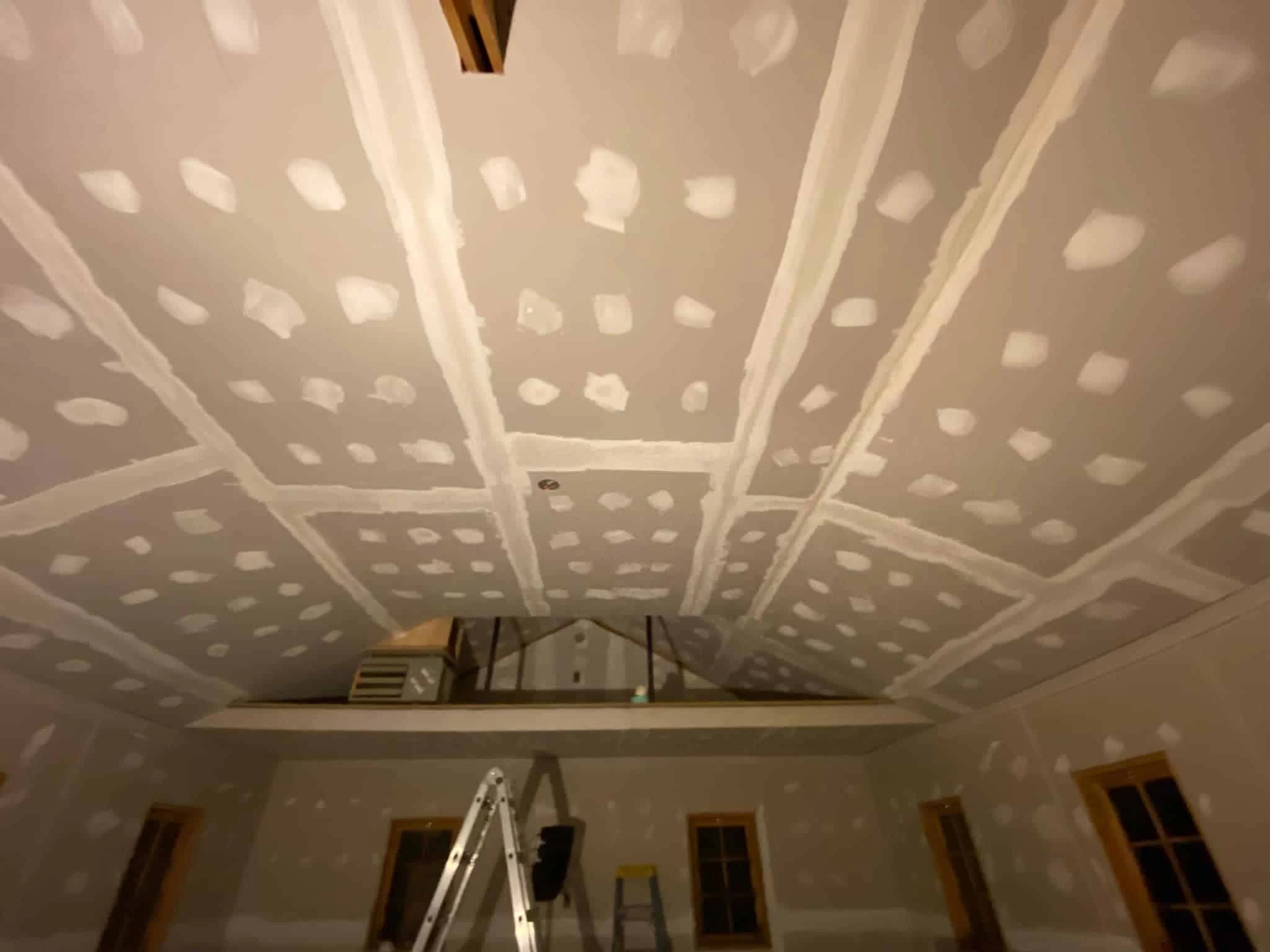 A room with unfinished drywall on the ceiling. Various tools and ladders are positioned around the room.