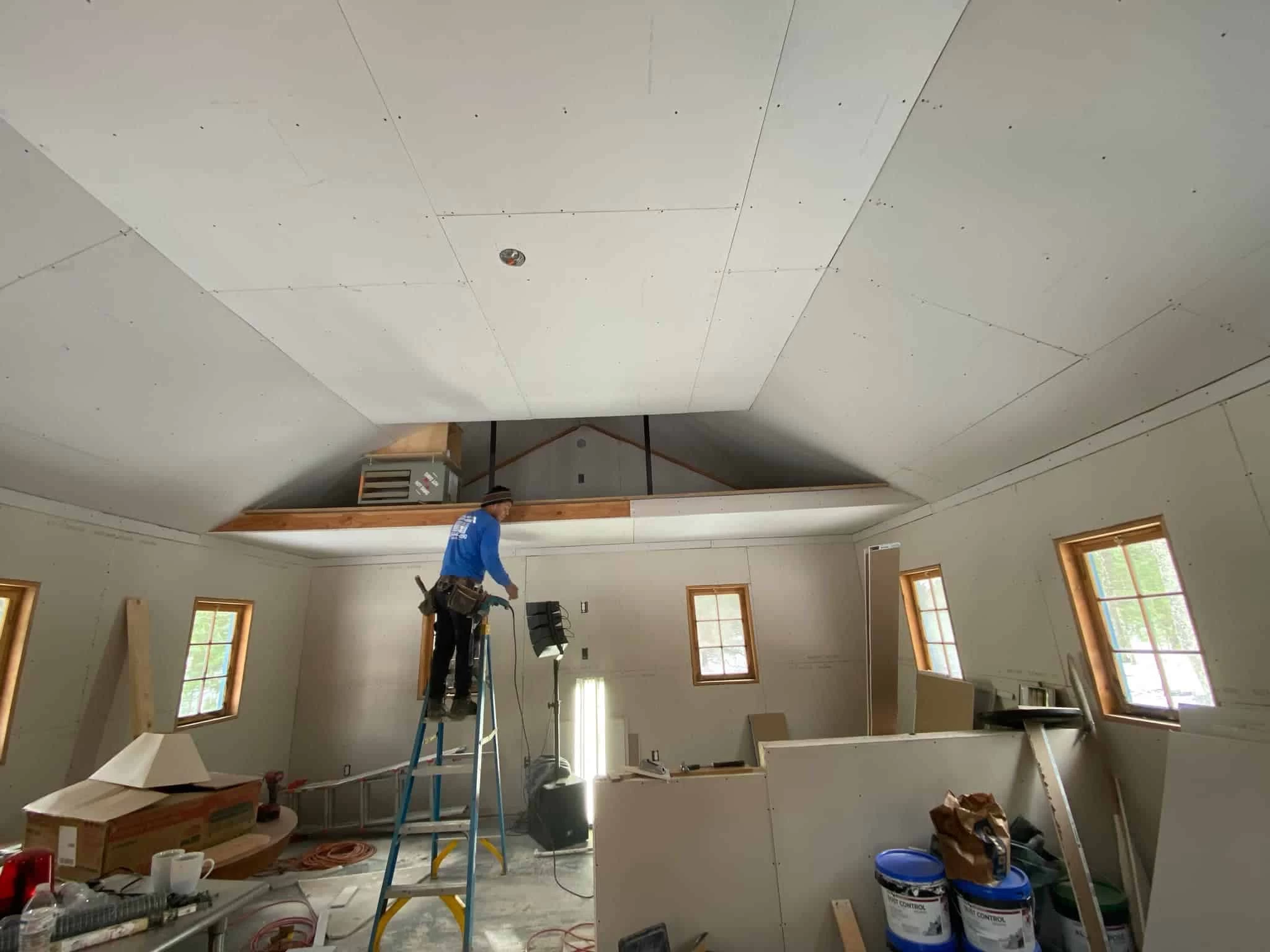 A construction worker stands on a ladder in a room under construction, focusing on the ceiling.