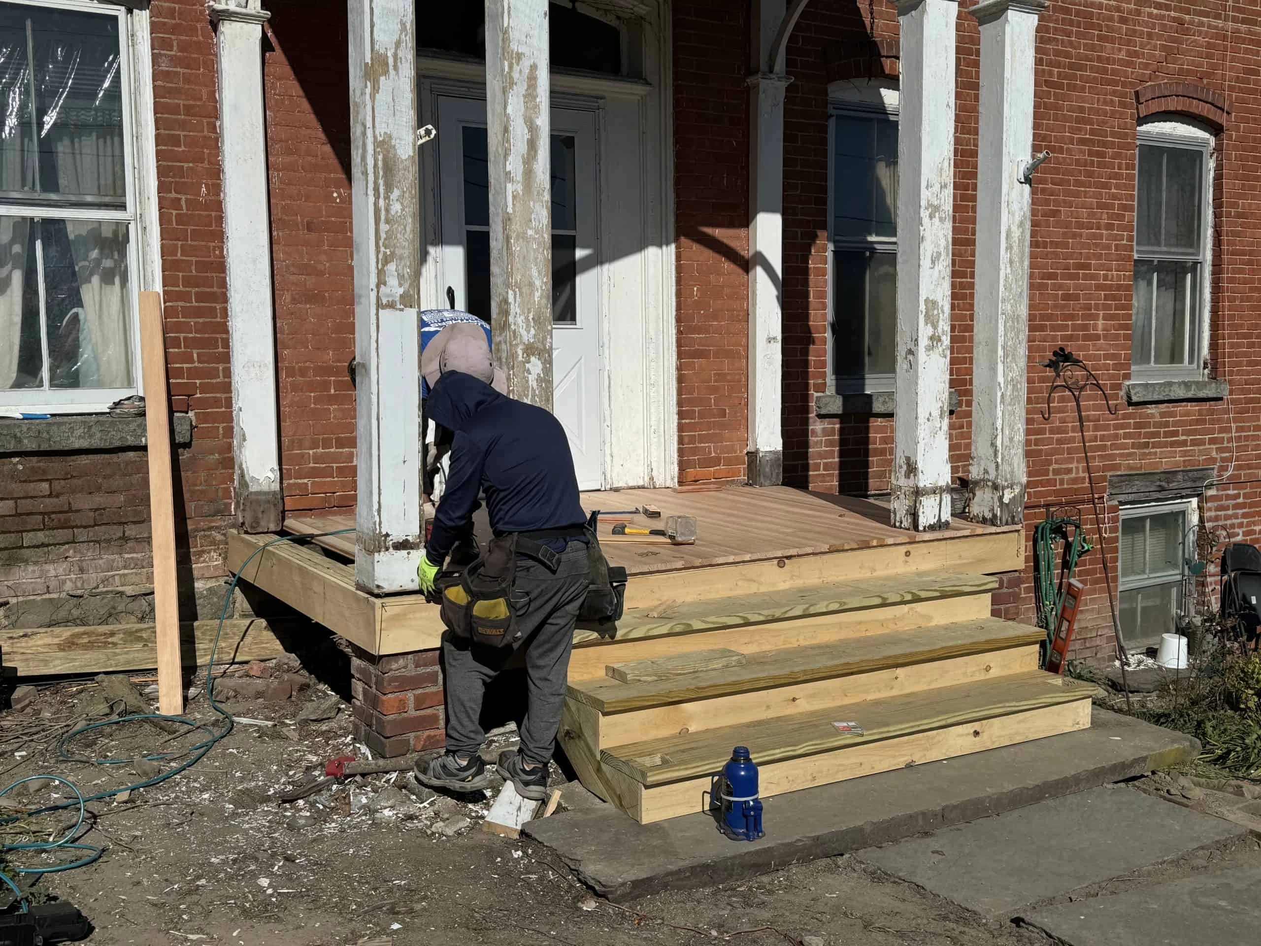 Person in a hoodie working on constructing a wooden porch in front of a red brick building.