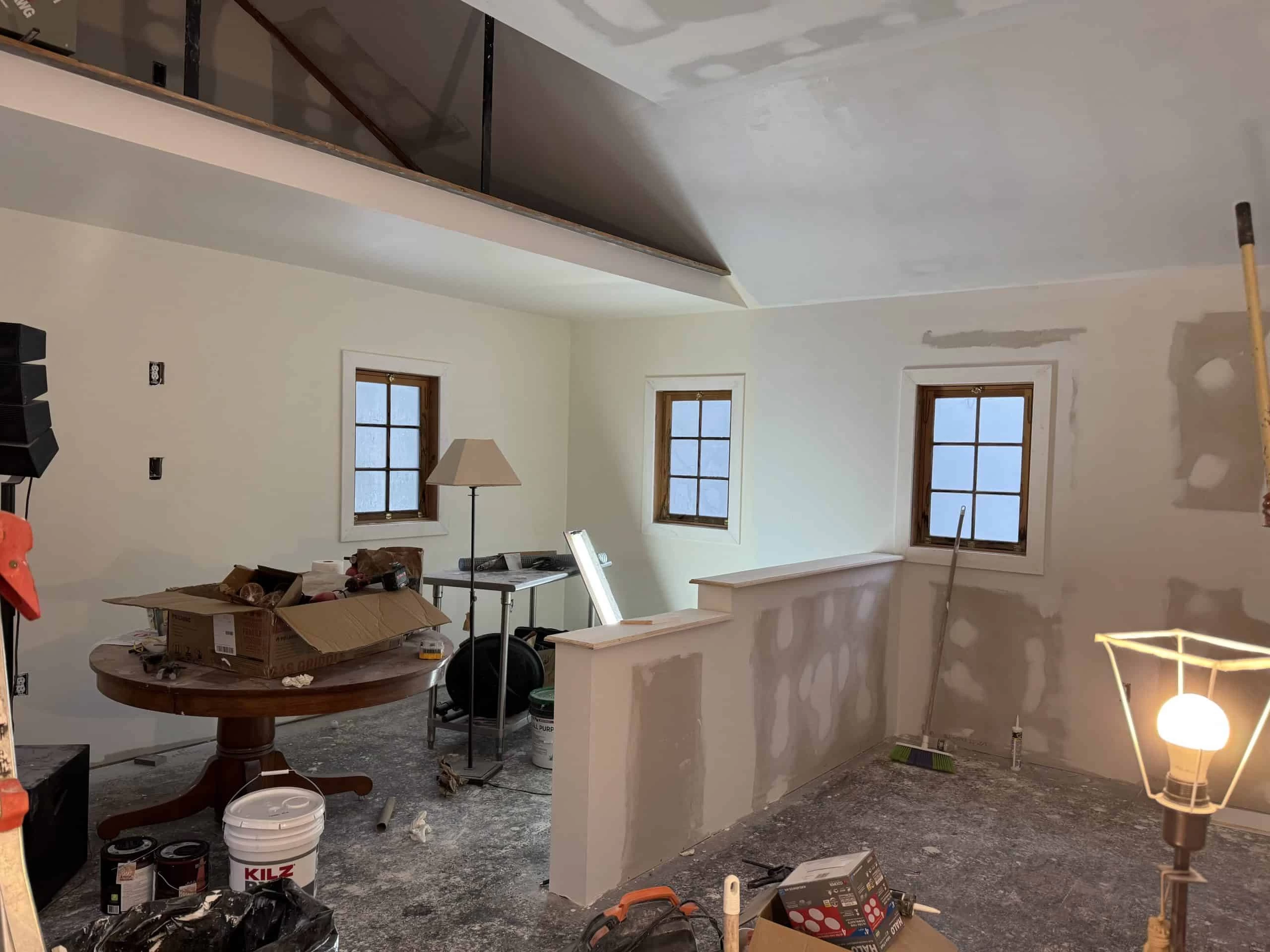 A partially renovated room with unfinished drywall, a round table covered with tools and boxes, and a lamp on.