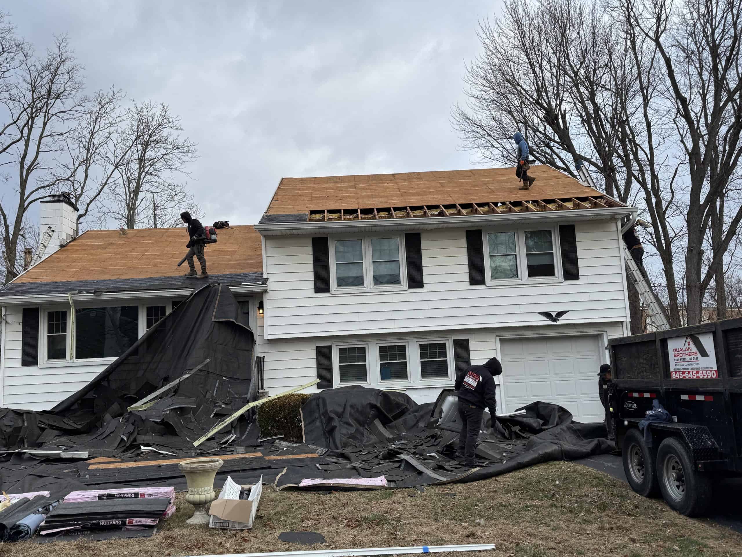 Workers are repairing the roof of a two-story white house with black shutters. One worker is on the roof laying new wooden boards, while others manage materials on the ground. The yard is covered with protective tarps, and there's a dumpster nearby.