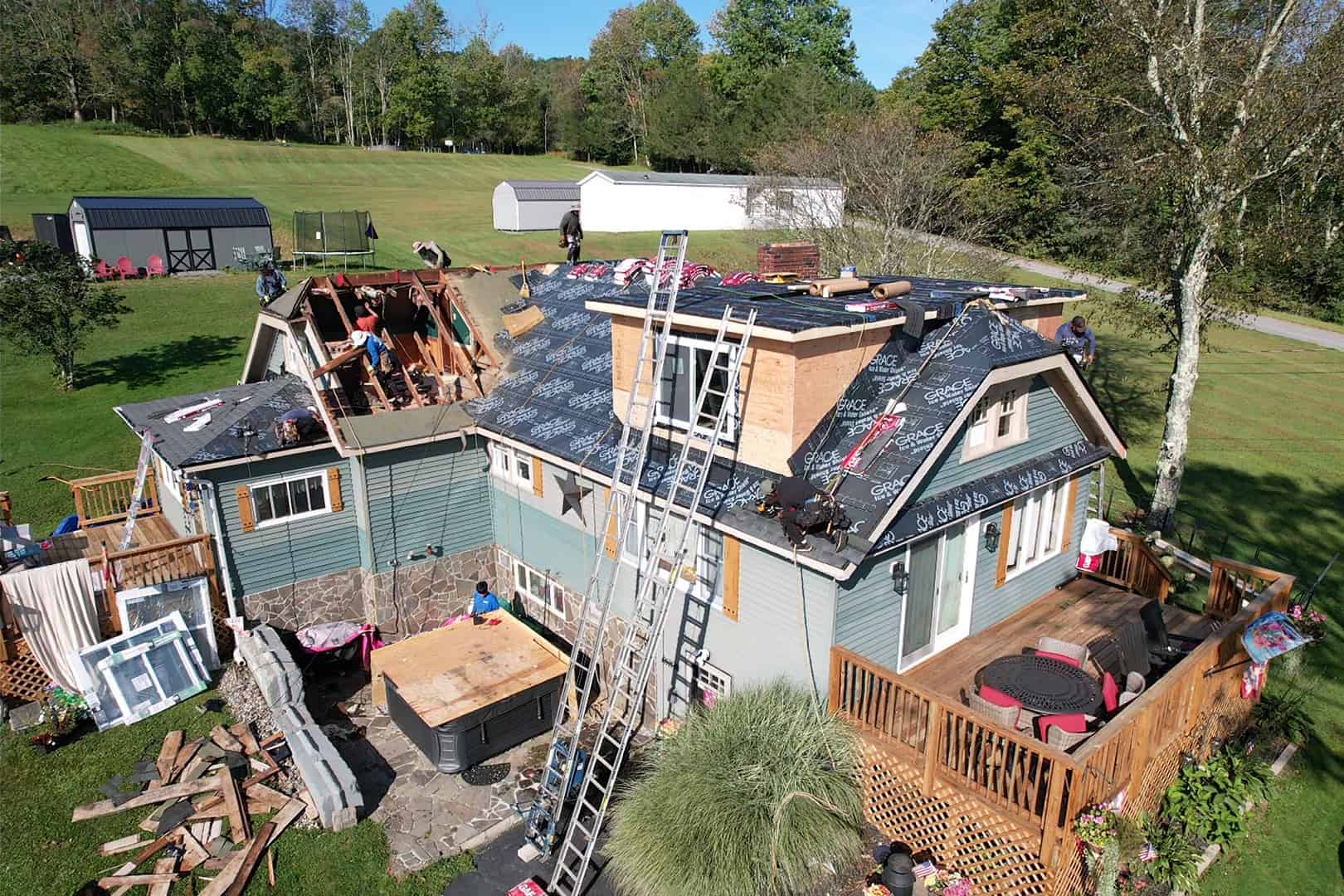 A house with a blue and gray exterior is under renovation. Several workers are on the roof, which is partially covered with black underlayment. Construction materials are scattered around, and two ladders lean against the house.