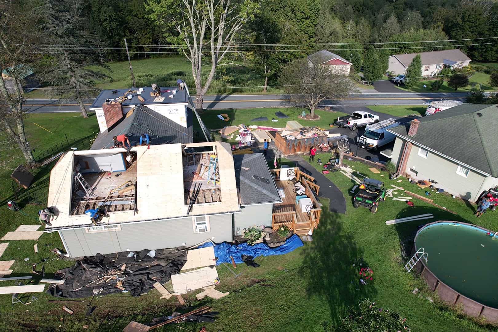 Aerial view of a house under renovation, with workers on the roof and construction materials scattered around. Nearby is a green backyard with a partially covered pool and several vehicles parked on the driveway. Surrounding are trees and neighboring houses.