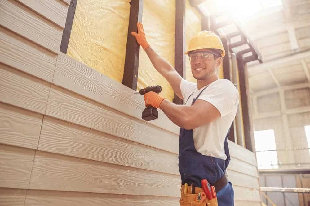 A construction worker in a hard hat and gloves uses a power drill on a wall with wood paneling.