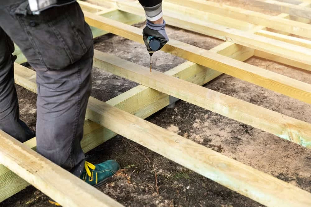 A person using a power drill to secure beams in a wooden deck framework. The person is wearing grey pants, gloves, and green shoes. The ground beneath is bare soil. Light shines on the wood, emphasizing the construction work in progress.
