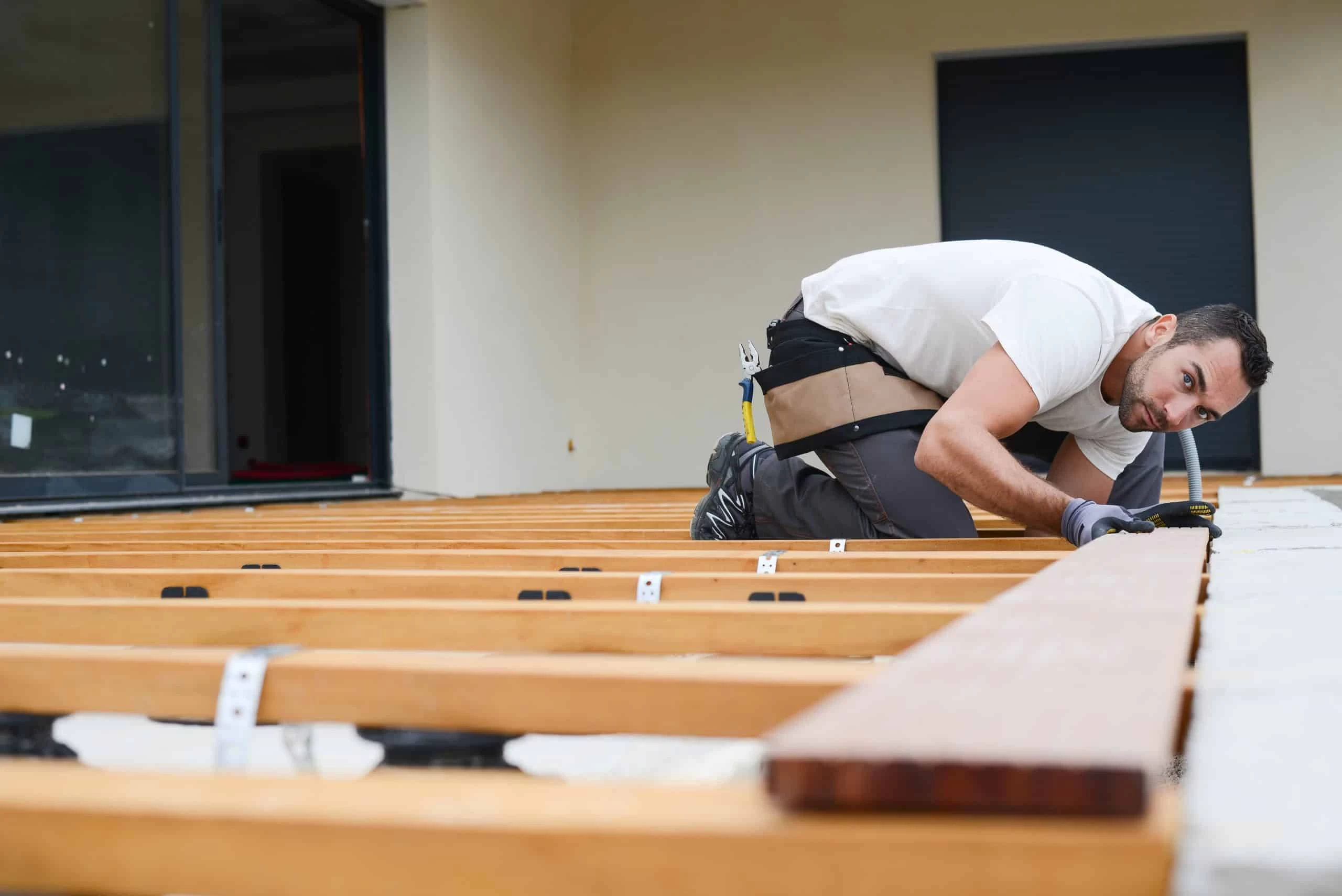 A person kneels, installing wooden beams for a deck on a patio. They wear a tool belt, gloves, and casual work attire.