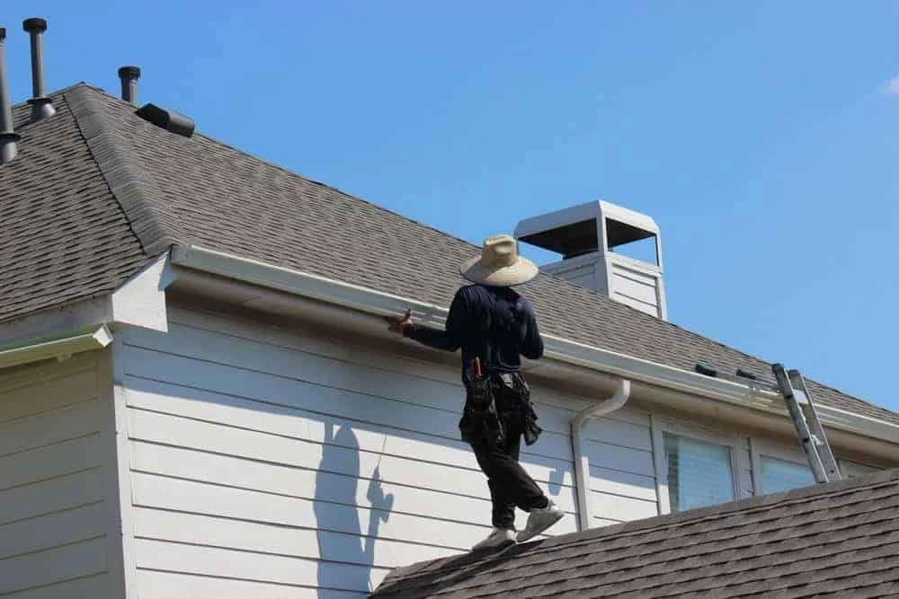 A person wearing a hat and tool belt stands on the roof of a house, cleaning or inspecting the gutters.