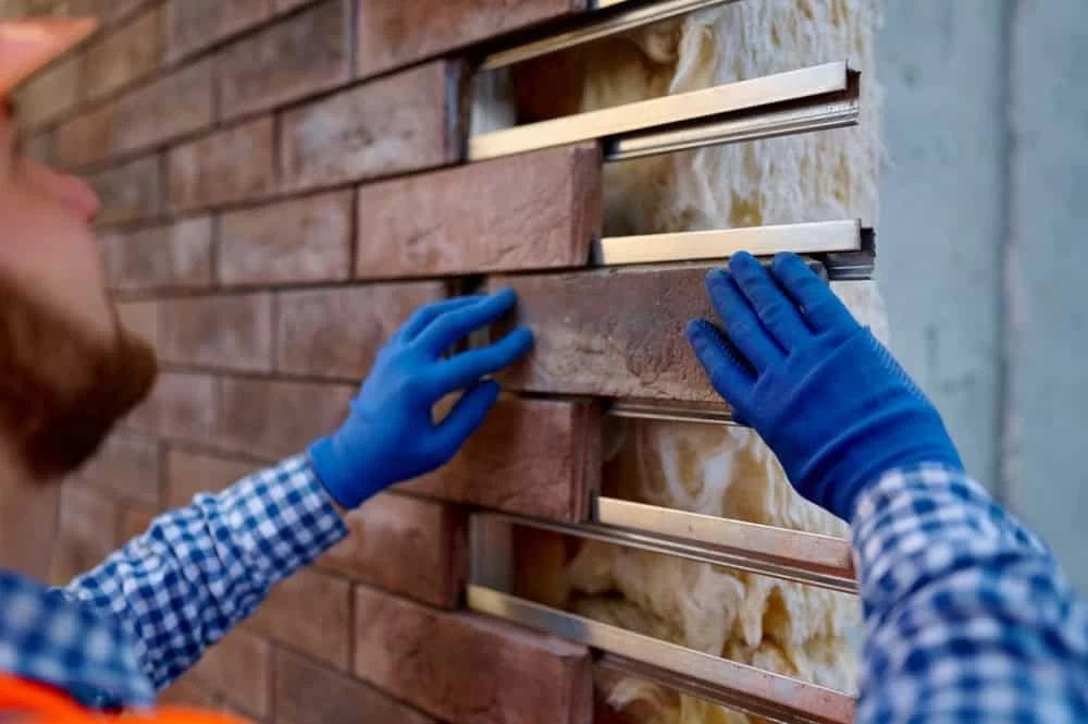 A construction worker in a blue checkered shirt and blue gloves installs brick siding on a wall.