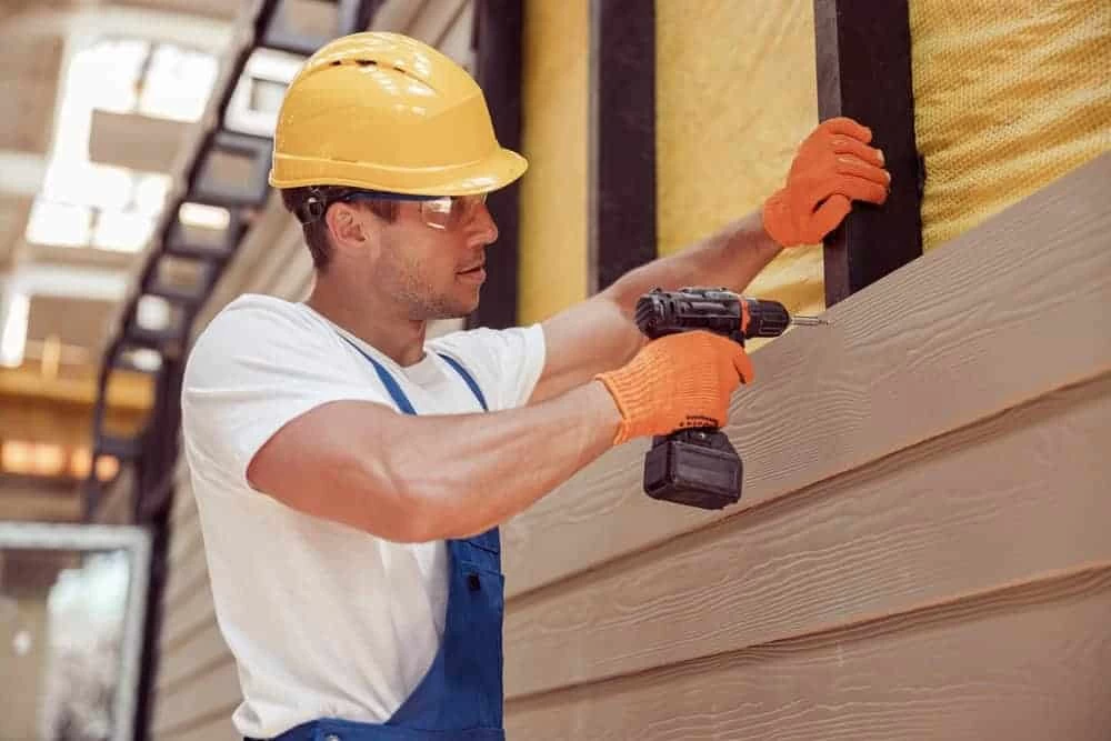 Construction worker in a yellow hard hat and orange gloves, using a power drill to fasten siding to a wall.