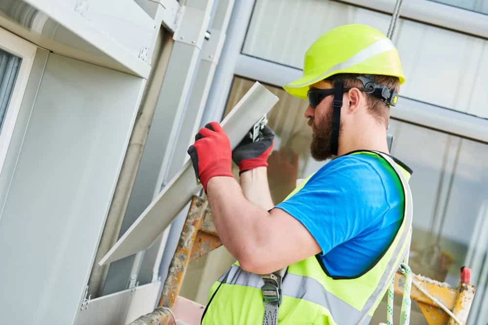 A construction worker in in Orange & Dutchess County wearing a yellow hard hat, sunglasses, green reflective vest, and red gloves is on scaffolding.