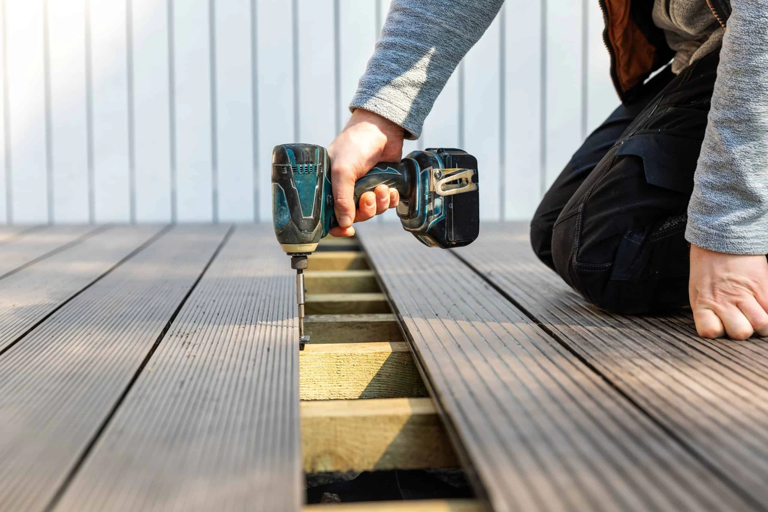 A person kneels while using a power drill to install decking boards. Their left hand steadies the board, while the right hand operates the drill. The background features light-colored wooden paneling.